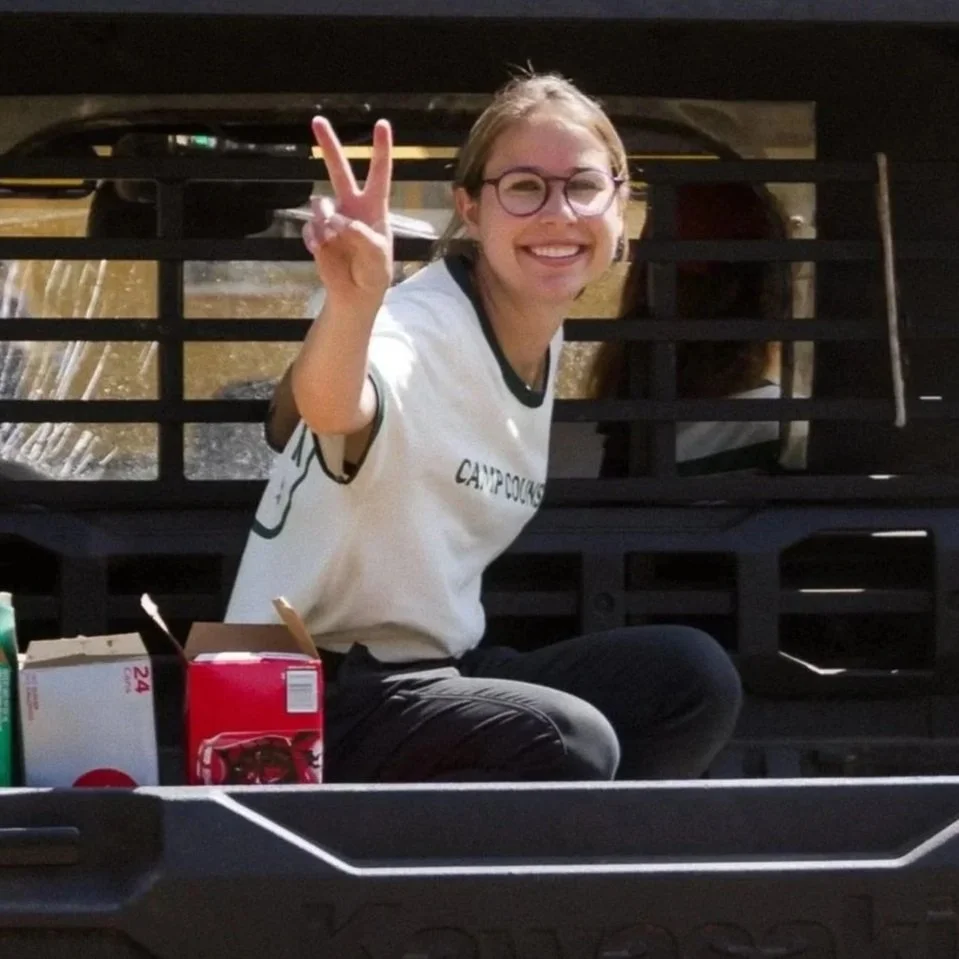 Young woman with glasses smiling and making a peace sign in a black truck bed, with a woman visible in the background and various snacks or drink boxes in the foreground.