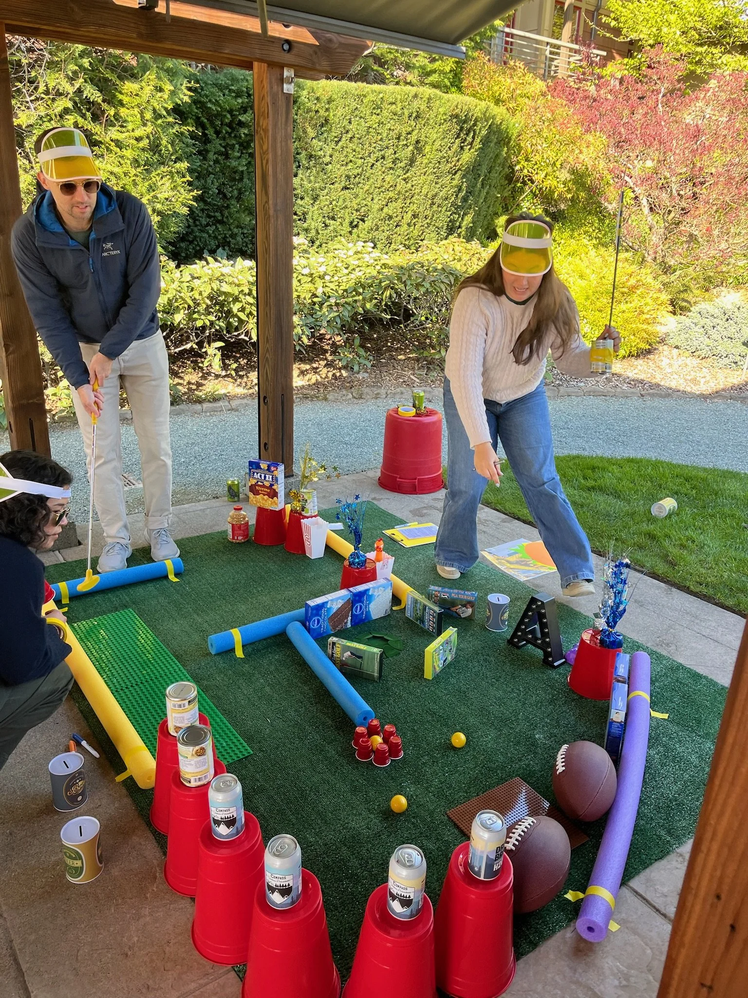 People playing a beer pong game outdoors under a wooden shelter with a backyard in the background. The area is decorated with plates, cans, and sports equipment like footballs and foam noodles.