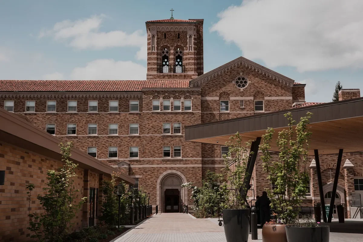 A large brick church with a tall bell tower, multiple windows, and a circular window, with a landscaped courtyard in front.