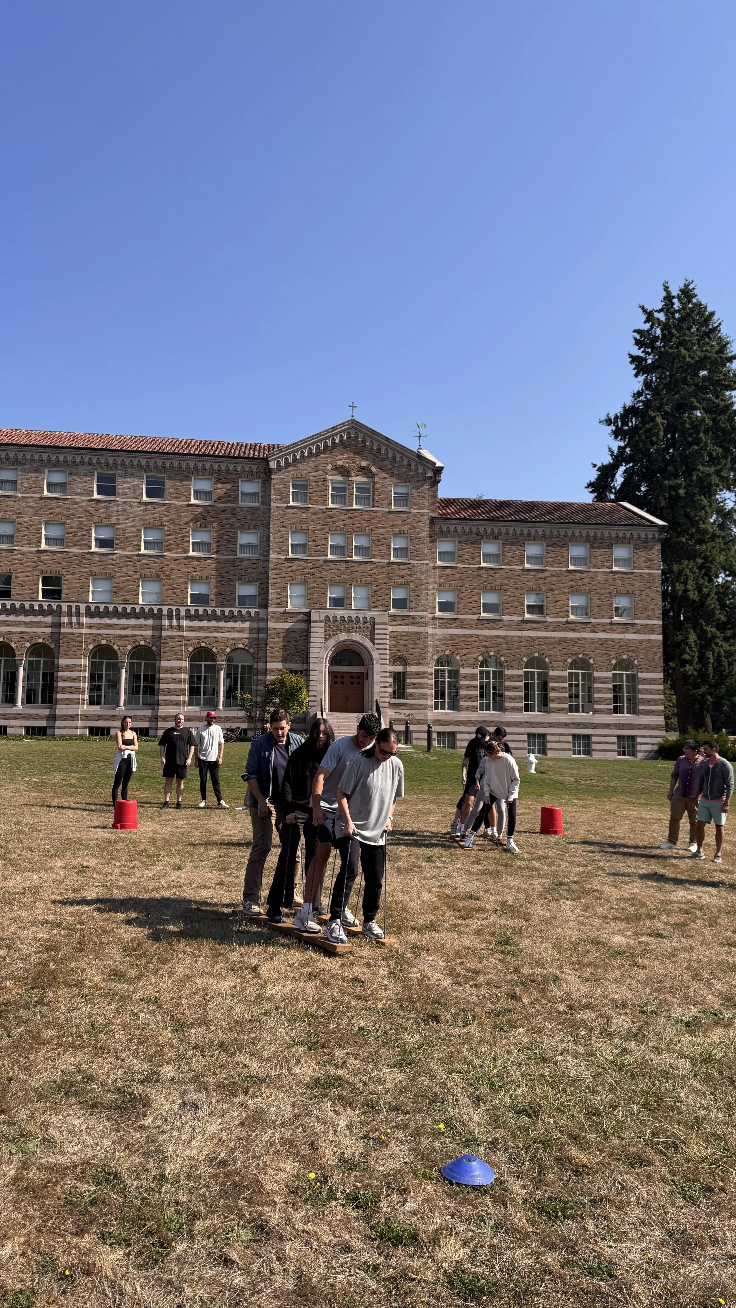 Students participating in an outdoor team-building activity on a grassy field in front of a historic brick building with arched windows on a sunny day.