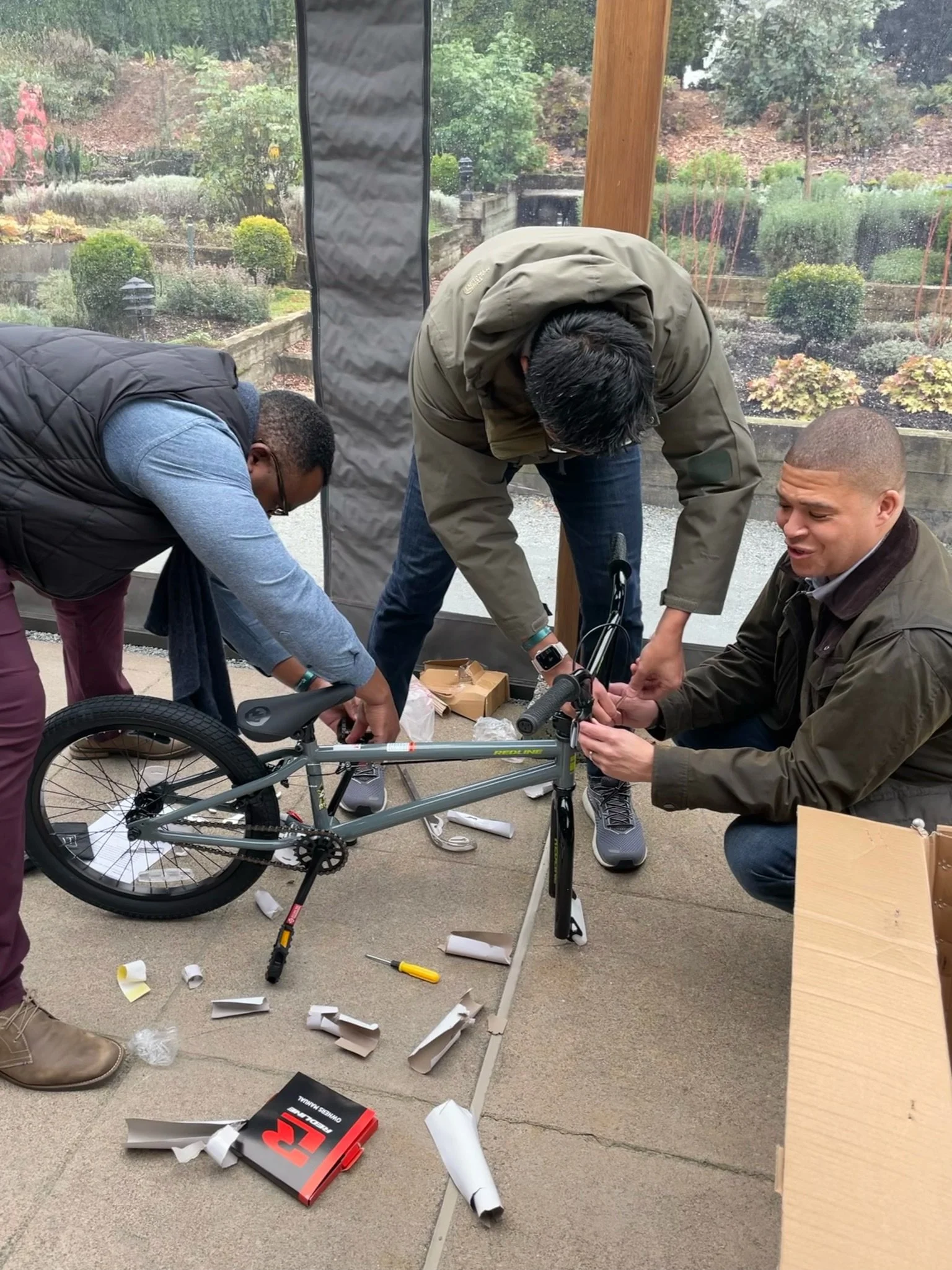 Three men assembling a gray bicycle on a concrete porch with torn packaging materials scattered around and a cardboard box. One man is kneeling and holding the handlebar, another is bent over working on the seat or frame, and the third is standing watching.
