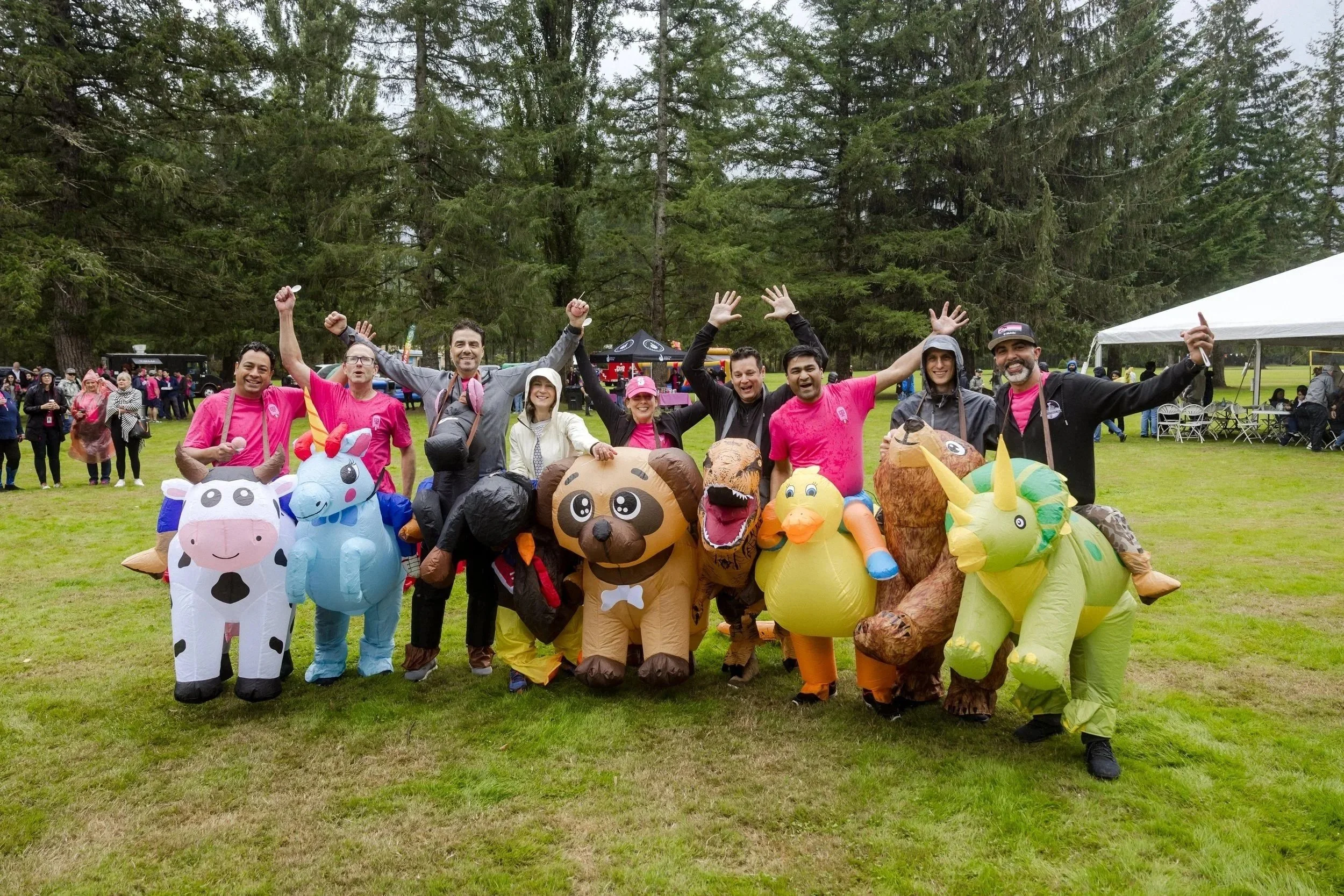 Group of people in animal-themed inflatable costumes celebrating outdoors in a park with trees and a tent in the background.