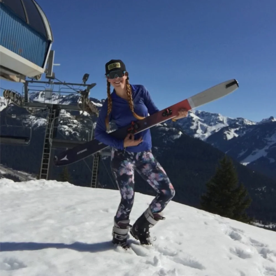 Woman smiling on snow-covered mountain, holding skis, wearing sunglasses, cap, colorful leggings, and ski boots, with ski lift and snow-capped mountains in the background.