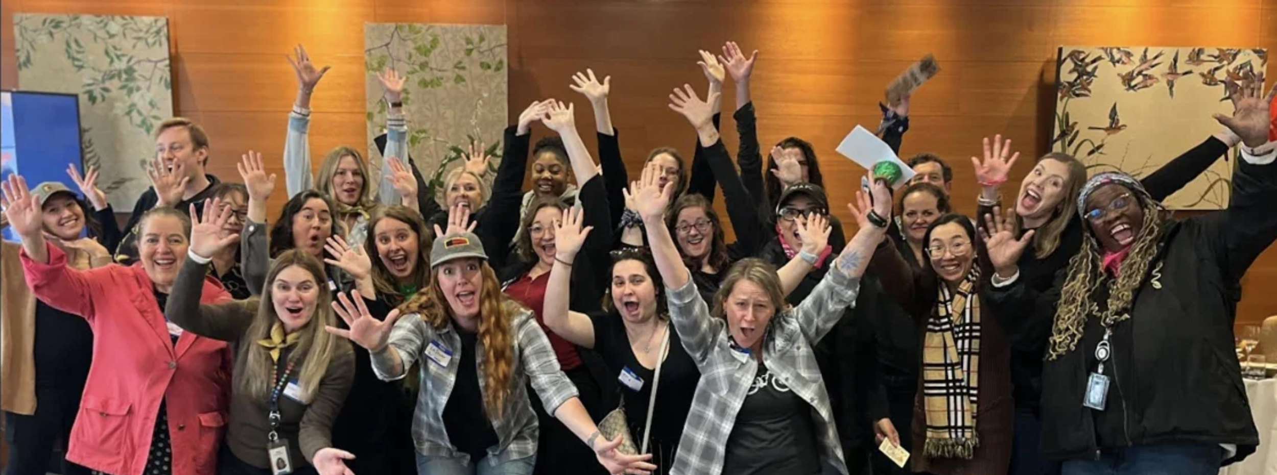 A large group of diverse women smiling and cheerfully waving in an indoor setting with a wooden wall and decorative artwork in the background.