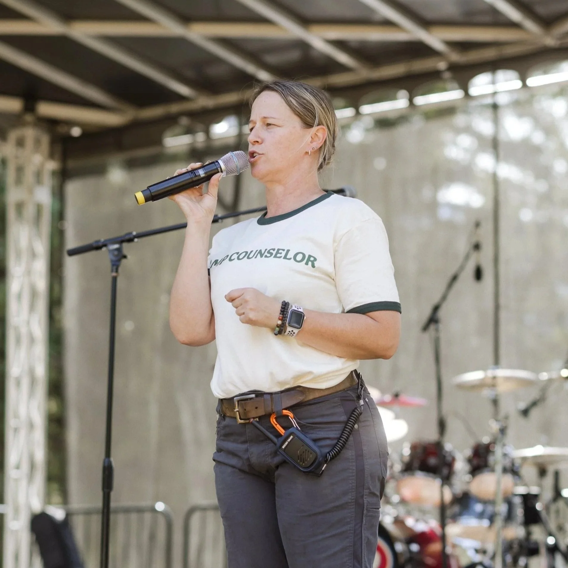 A woman speaking into a microphone on an outdoor stage, wearing a "VMP COUNSELOR" t-shirt, black pants, and a smartwatch.