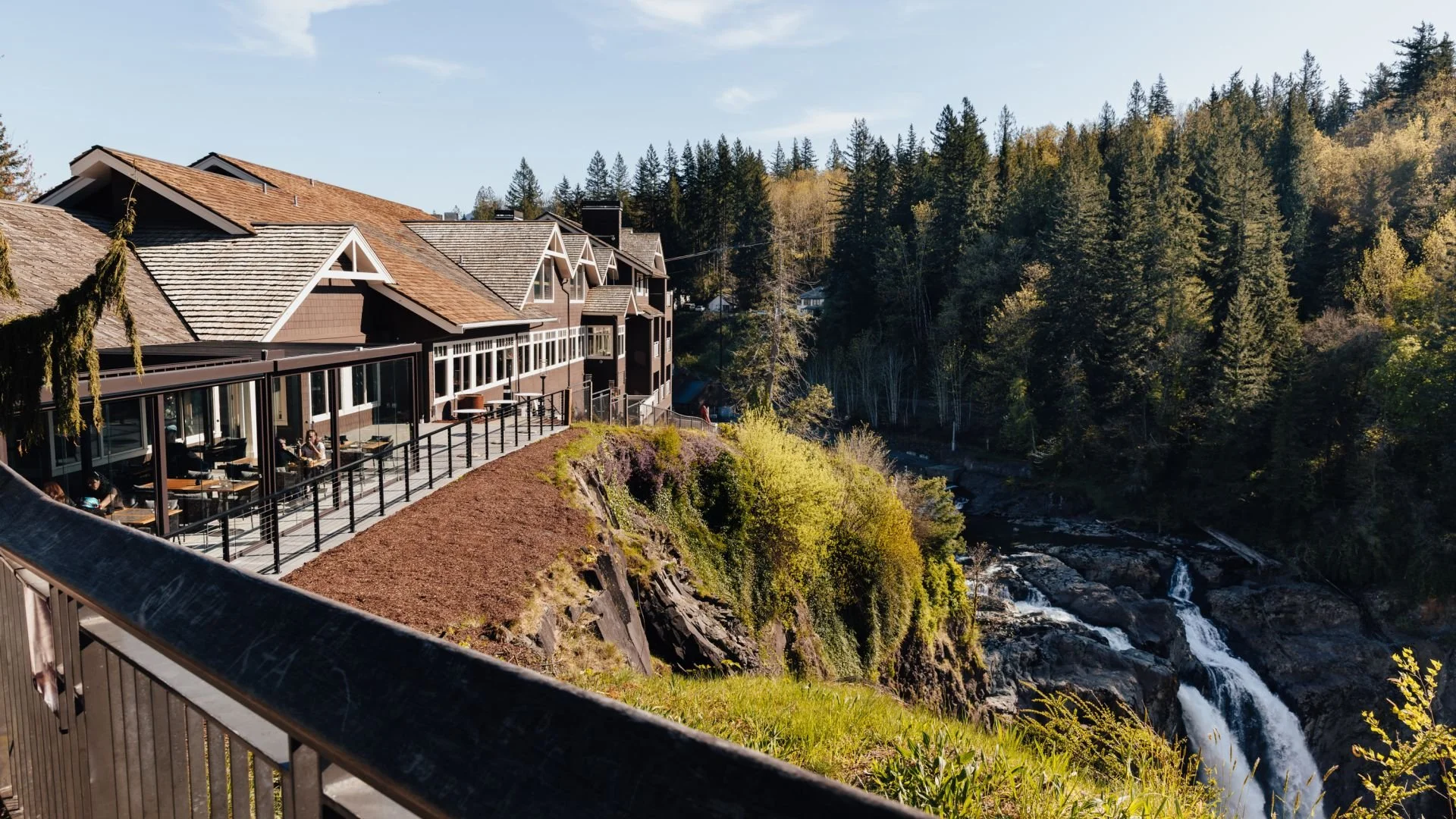 A scenic view of a building with large glass windows, an outdoor patio, and a wooden railing overlooking a waterfall and a forested hillside during daytime.