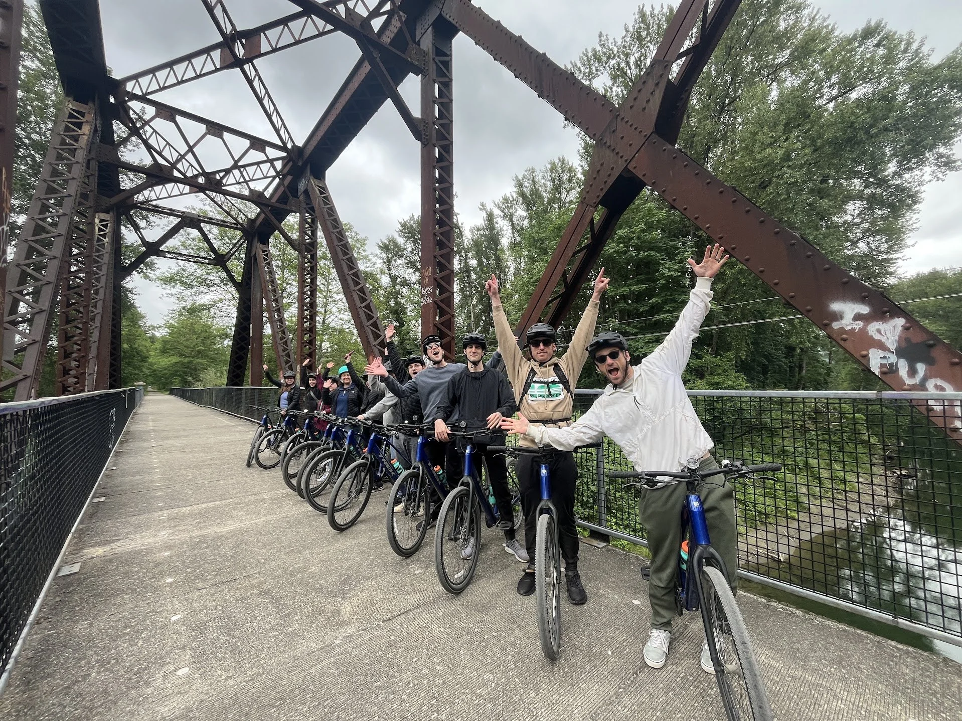 A group of people with bicycles on a bridge under a metal structure during daytime, some raising their arms and smiling.