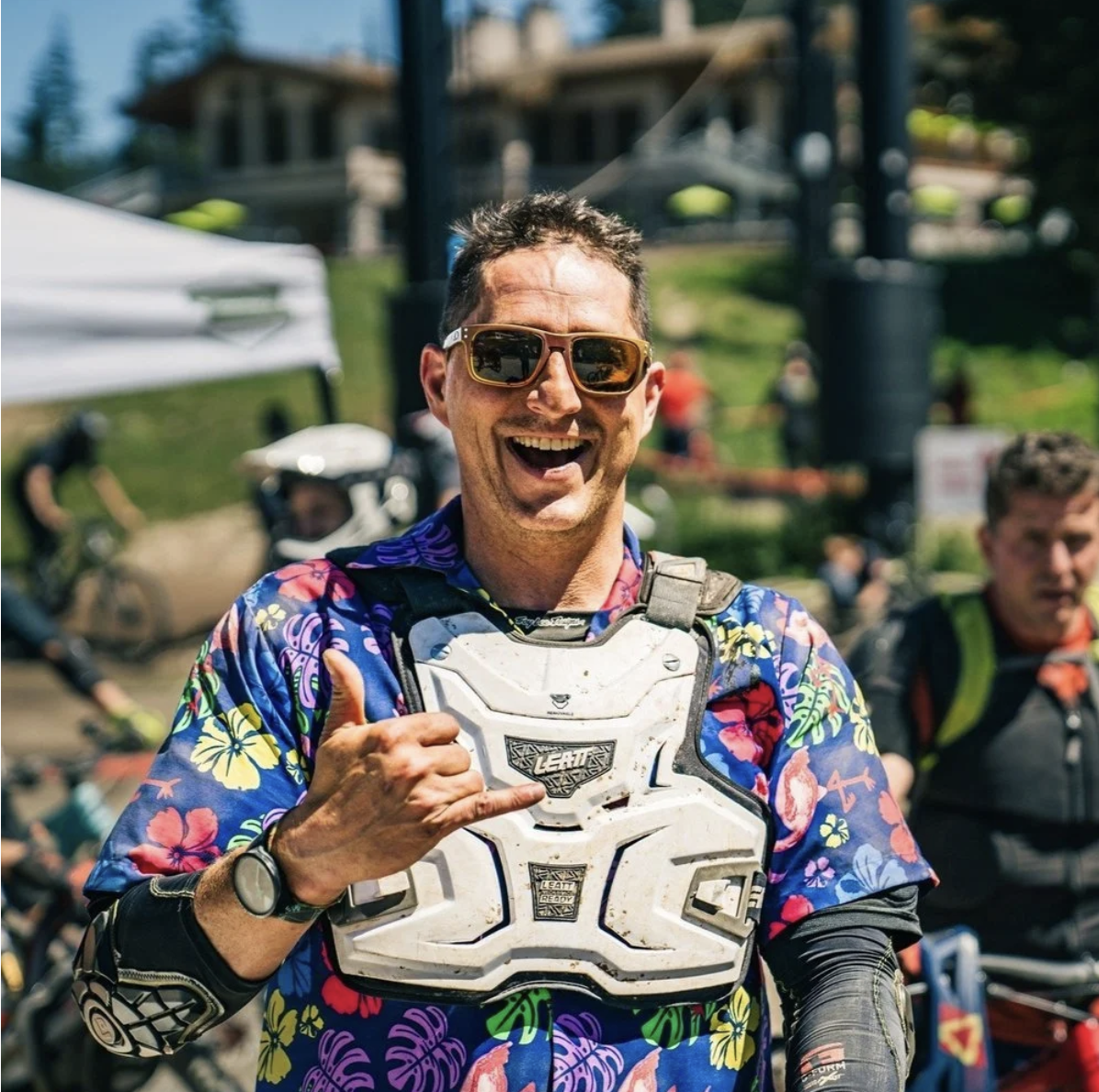 Johnny smiling and wearing sunglasses, a colorful Hawaiian shirt, and protective gear, giving a thumbs-up gesture at an outdoor event on a sunny day with mountain biking activity in the background.