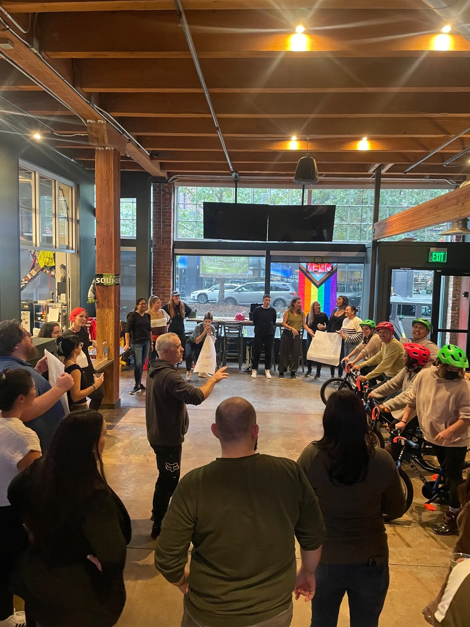 A group of people are gathered indoors, some with bikes and helmets, listening to a person speaking in the center. Others are standing around or sitting, with some holding signs. The room has large windows, a wooden ceiling, and a welcoming atmosphere.