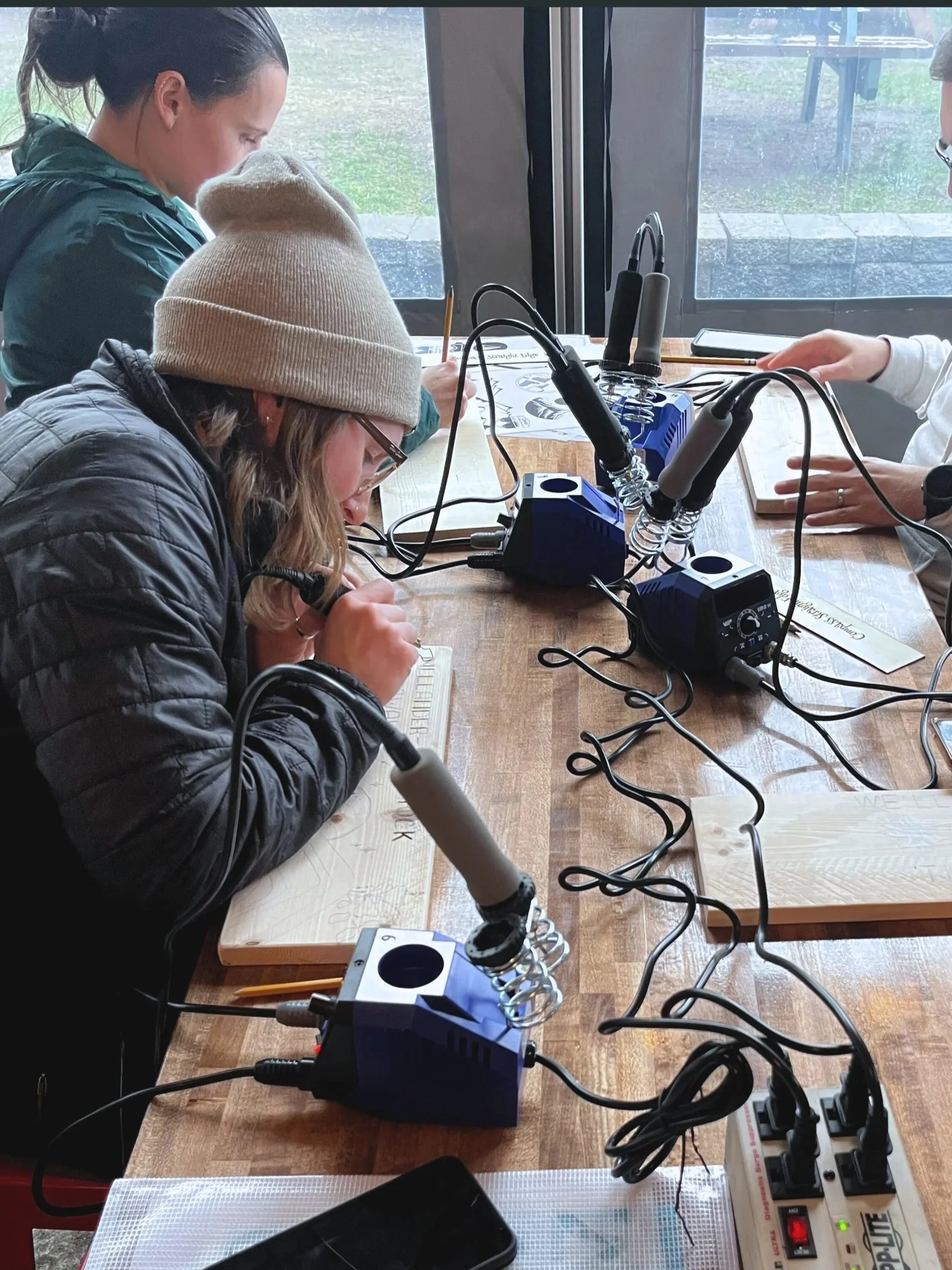 People working on soldering projects around a wooden table with soldering irons and power supplies.
