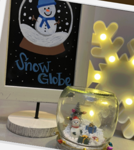 A snow globe with a snowman figurine inside, surrounded by pretend snow and small decorative items. In the background, there is a sign with a snowman and the words "Snow Globe" and a yellow decorative snowflake light.