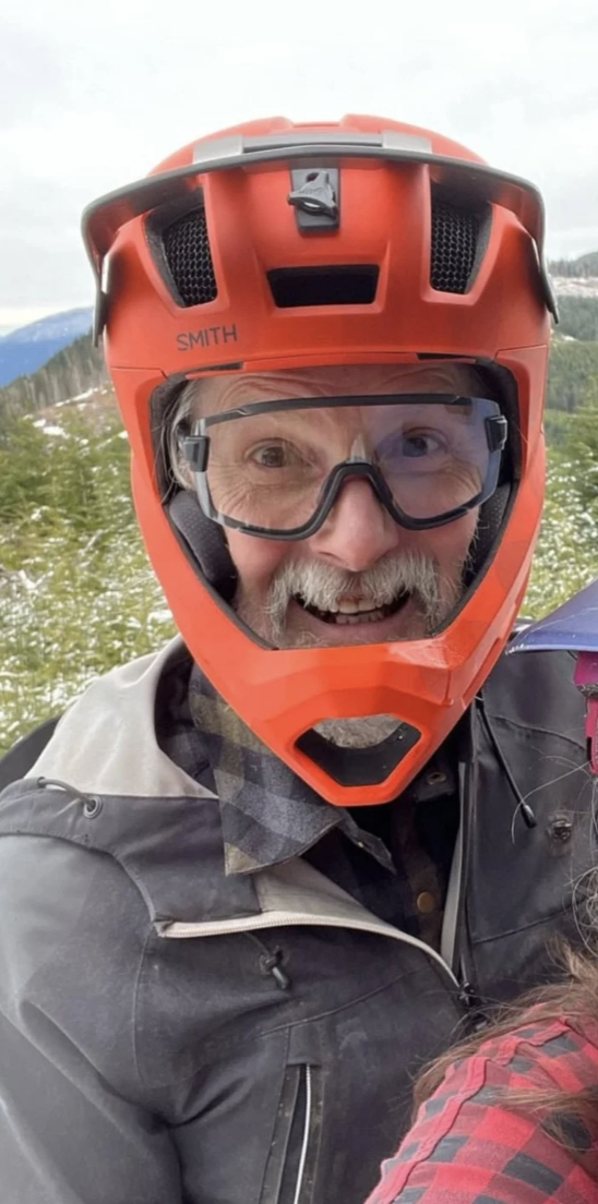 Man wearing a red motorcycle helmet and goggles, smiling outdoors with mountains and greenery in the background.