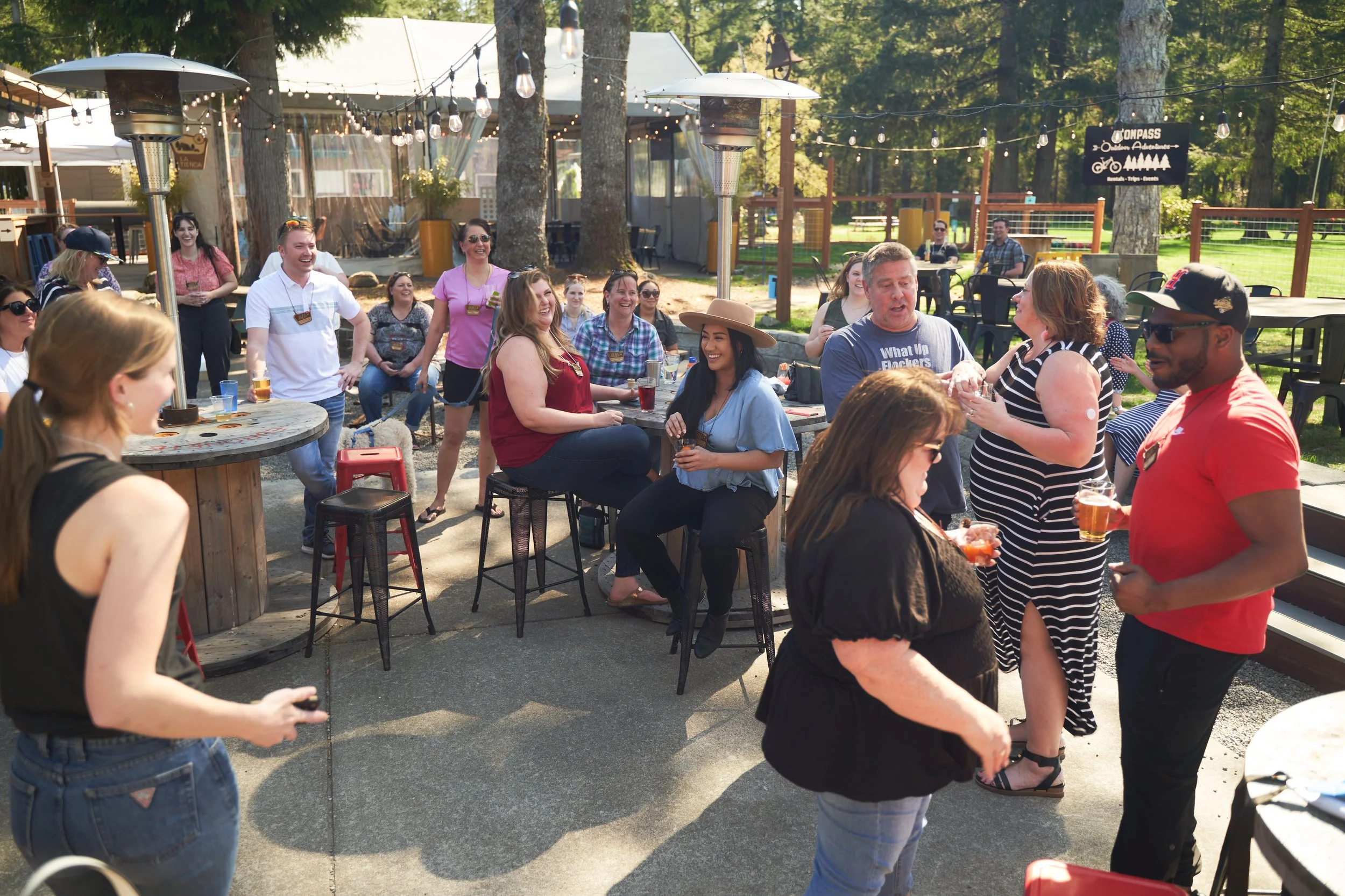 A group of people at an outdoor gathering, socializing and enjoying drinks, with string lights overhead and outdoor heaters present.