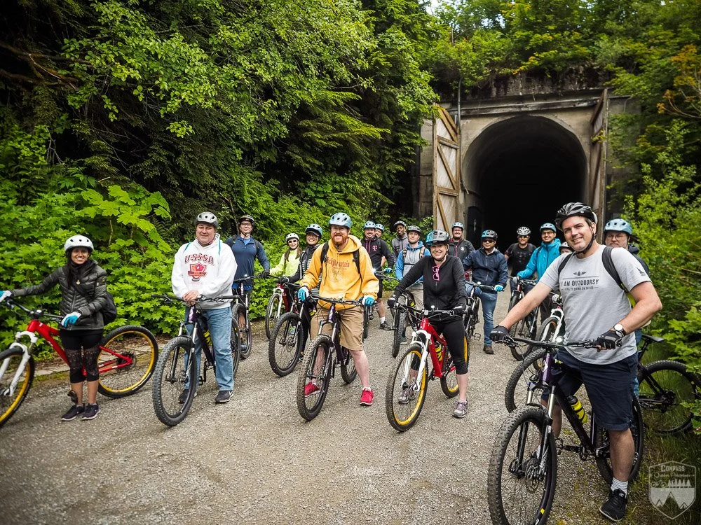 Group of men and women in cycling gear and helmets standing with bikes in front of a tunnel entrance surrounded by green foliage.