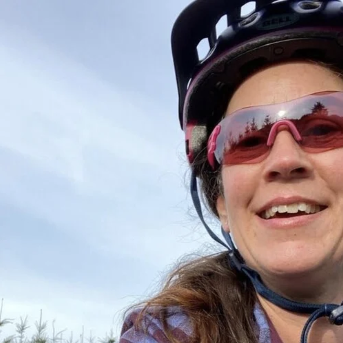 Close-up of a woman wearing a helmet and red sunglasses, smiling outdoors with a blue sky in the background.