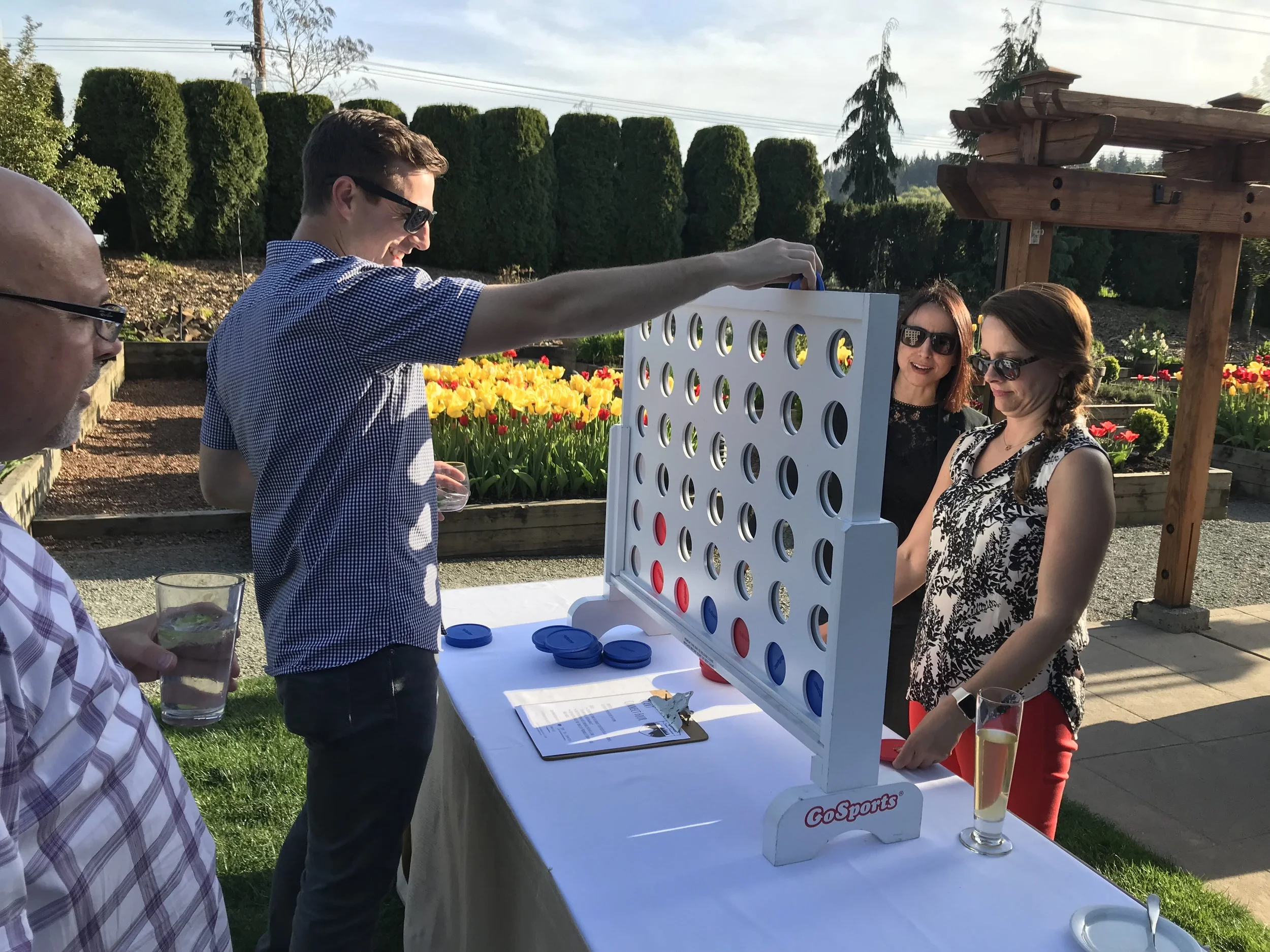 People playing a giant Connect Four game outdoors at a garden party, with tulip beds and trees in the background.