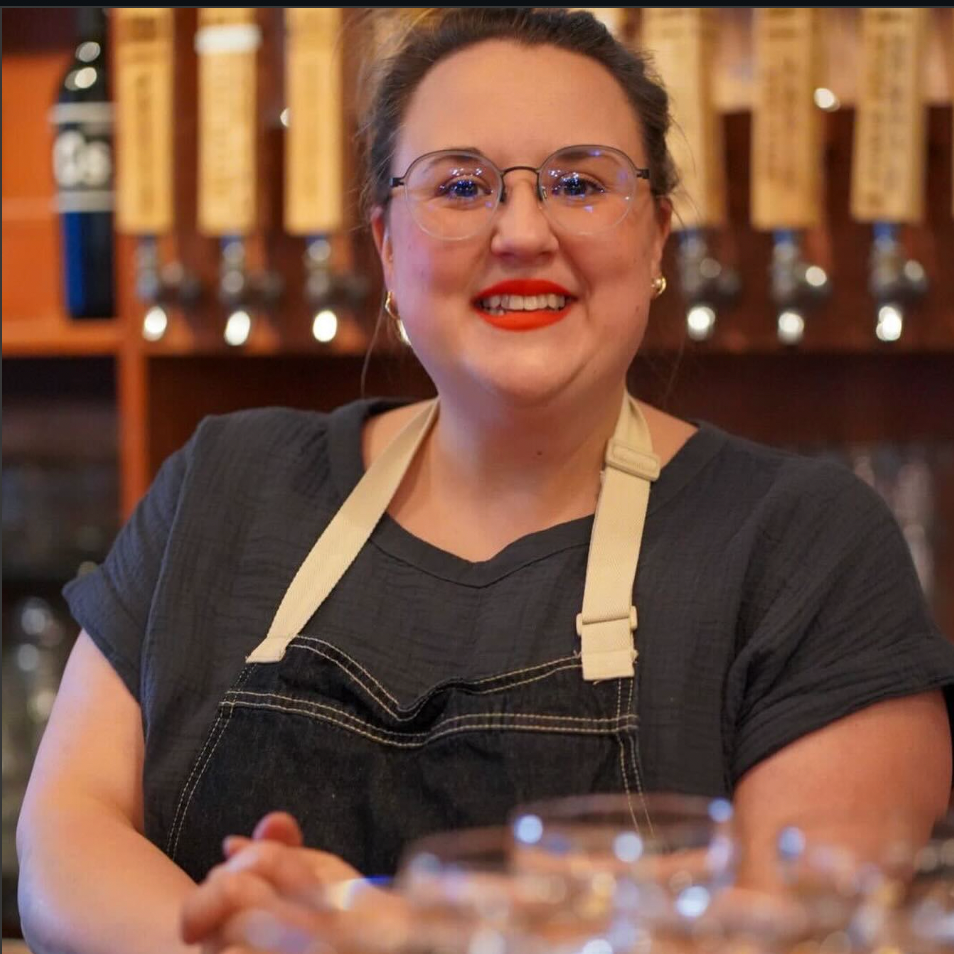 A smiling woman wearing glasses, bright red lipstick, a black shirt, and a beige apron, standing behind a bar with various taps in the background.