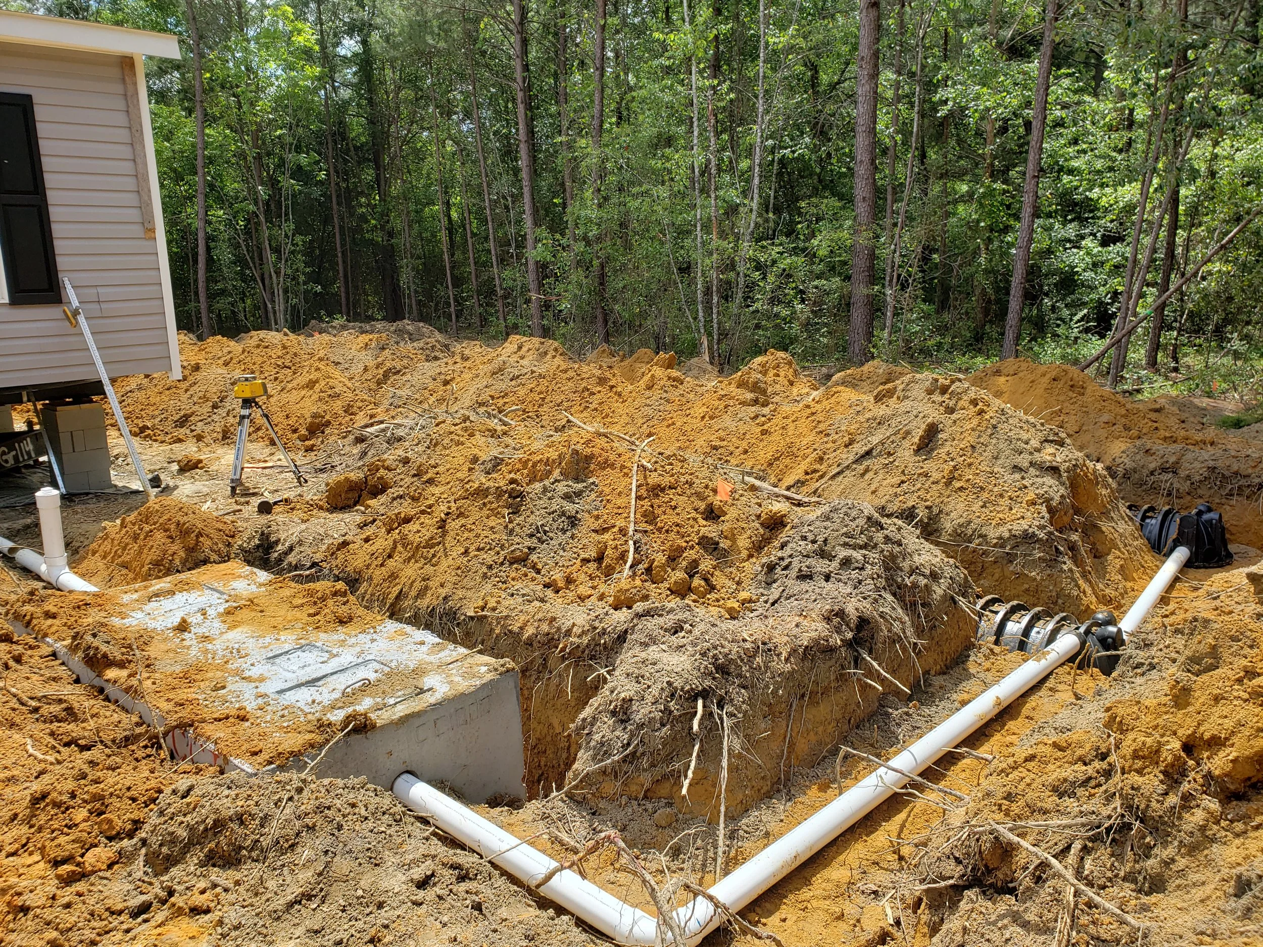 Construction site with exposed soil, pipes, and equipment next to a house, surrounded by trees.