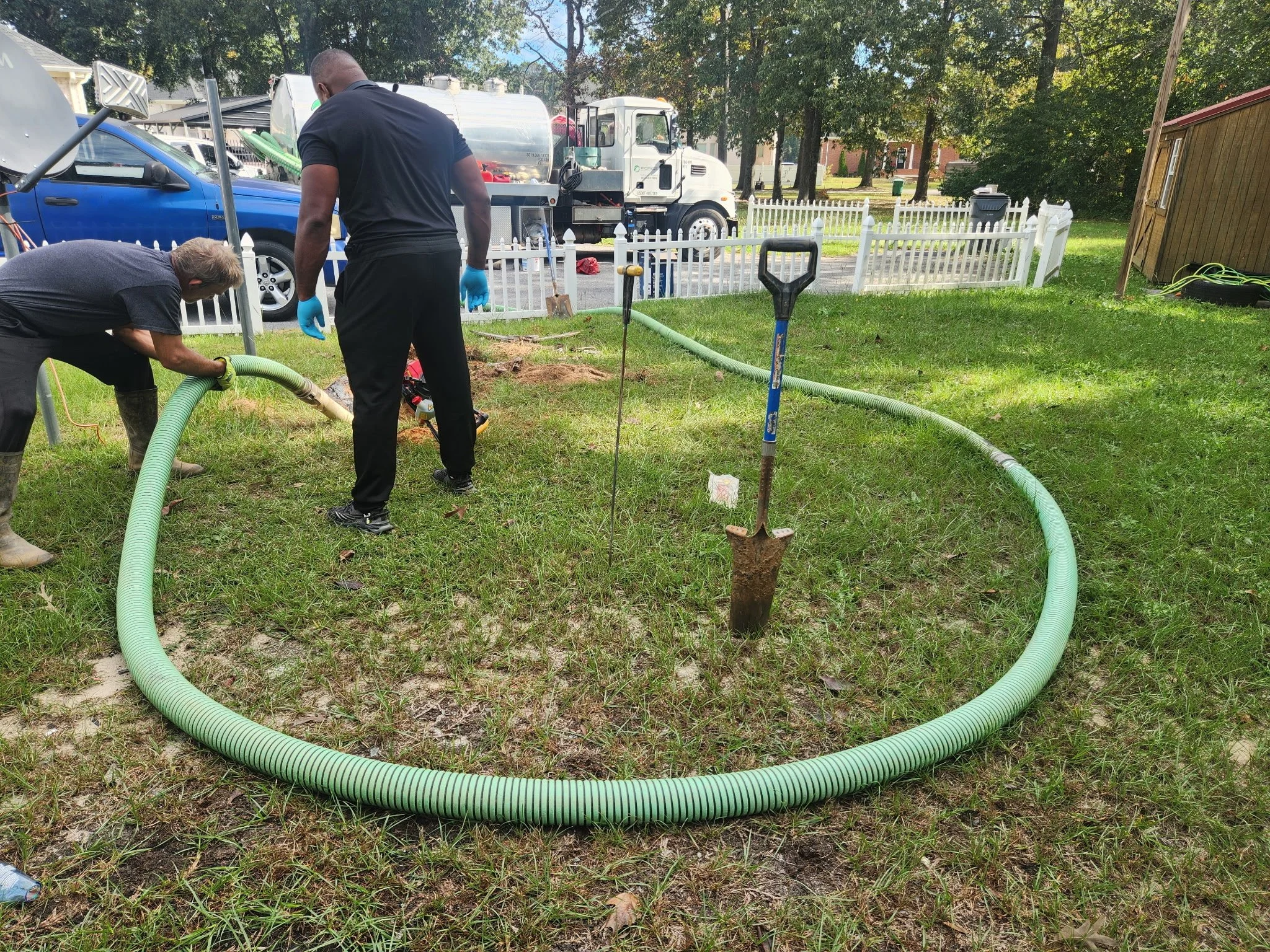 Two workers installing a large green corrugated pipe in a grassy yard, with tools and a shovel nearby, and vehicles and a white fence in the background.