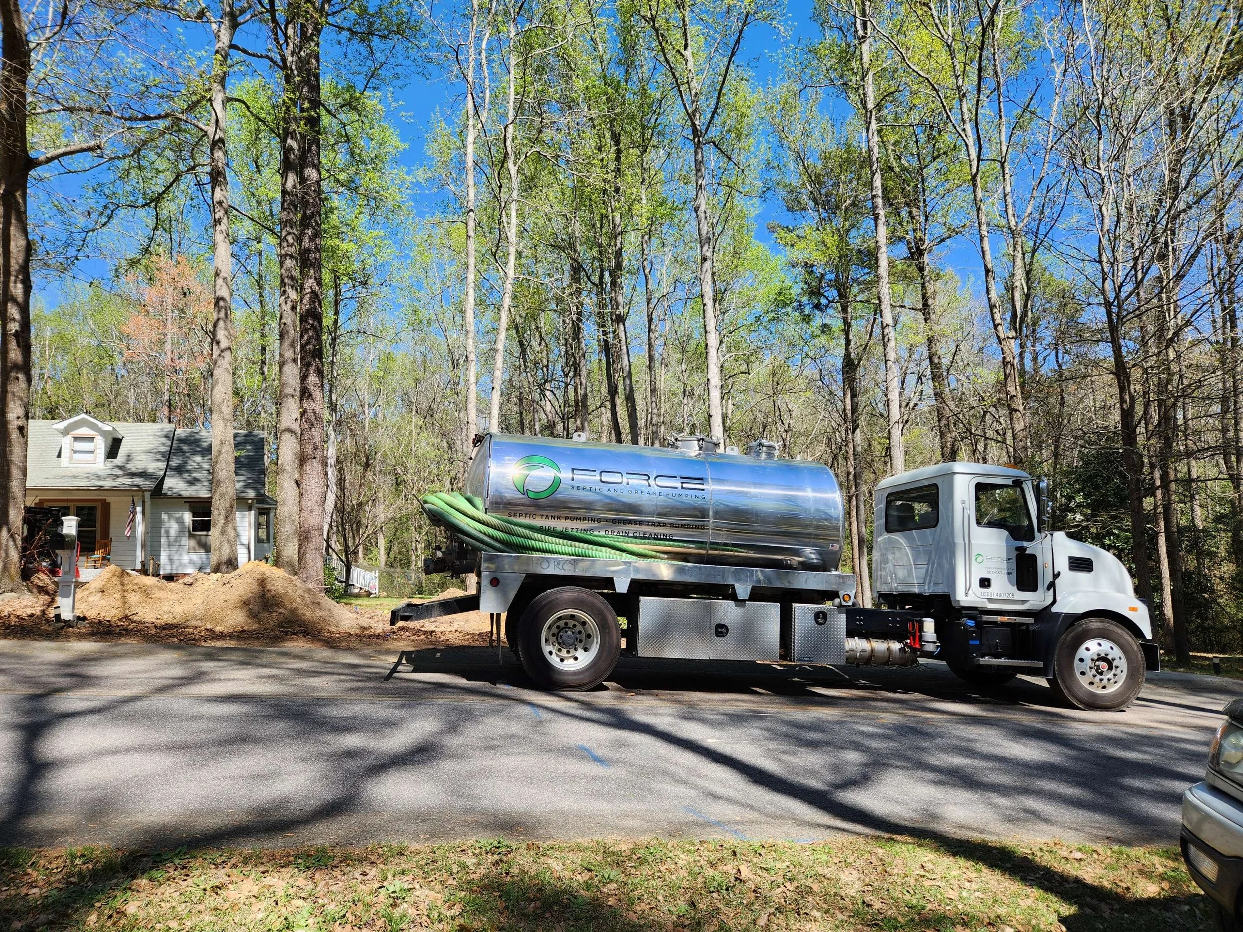 A septic tank pumping truck parked on a paved road in front of a wooded area with a house in the background. The truck has a large silver cylindrical tank with hoses attached.