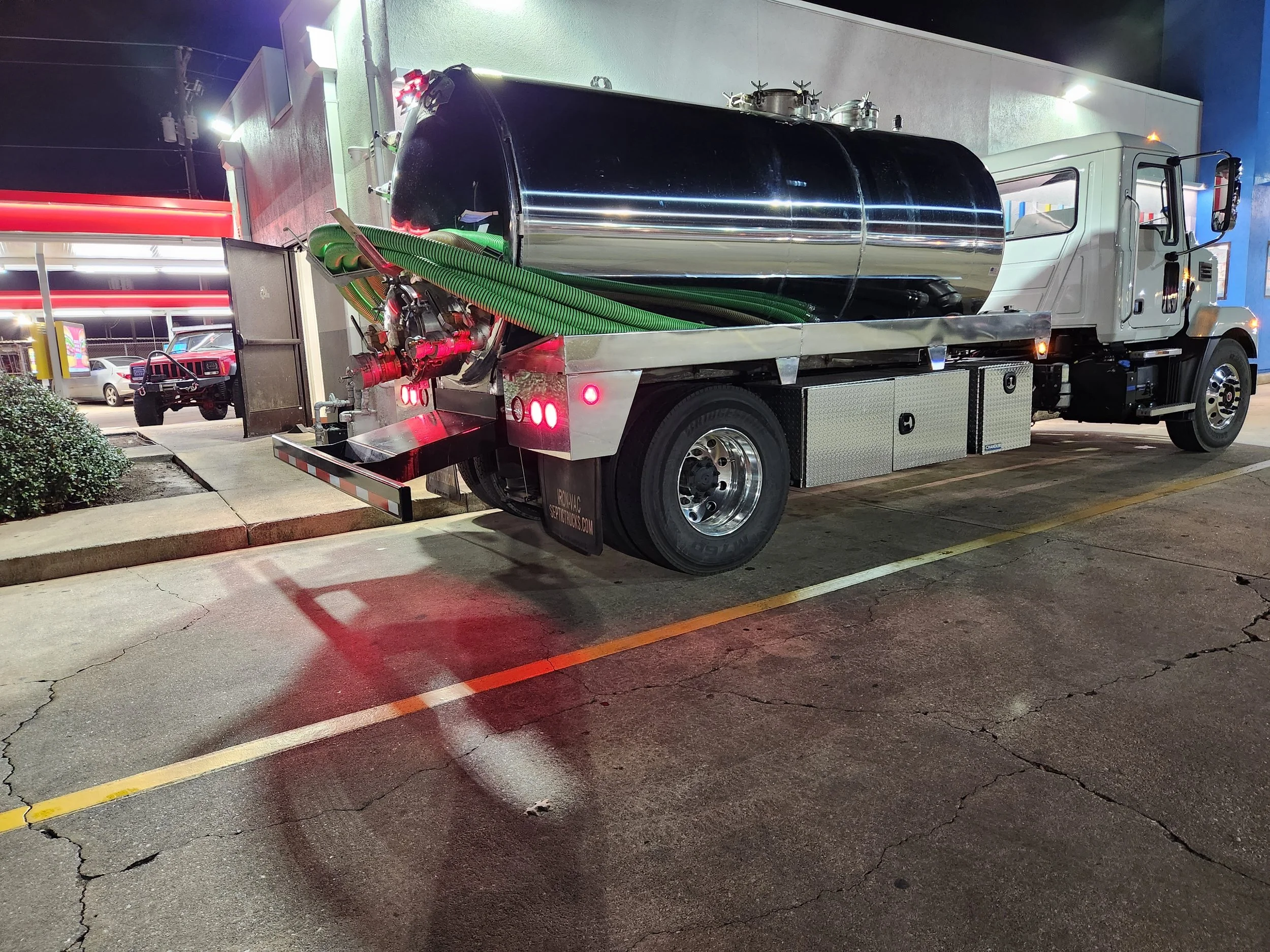 A large white fuel or water tanker truck with a shiny metal tank is parked in a parking lot at night. The truck has green hoses connected at the rear, with some red equipment and lights visible.