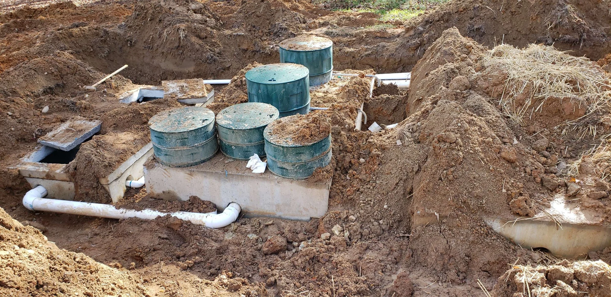 Underground plumbing installation with several blue sewer or drain pipes and white PVC pipes in a dirt trench, surrounded by dirt mounds and some dry grass.