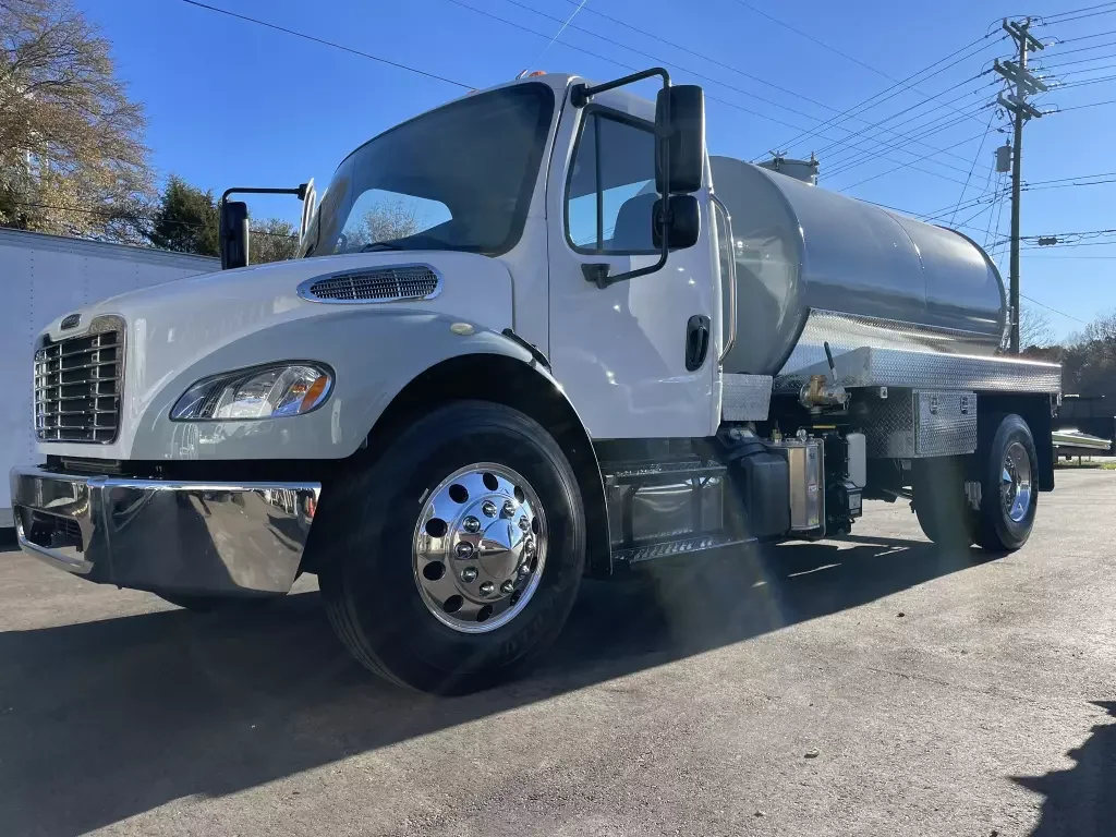 A white tanker truck parked on a paved surface with electrical poles in the background, blue sky, and some trees.
