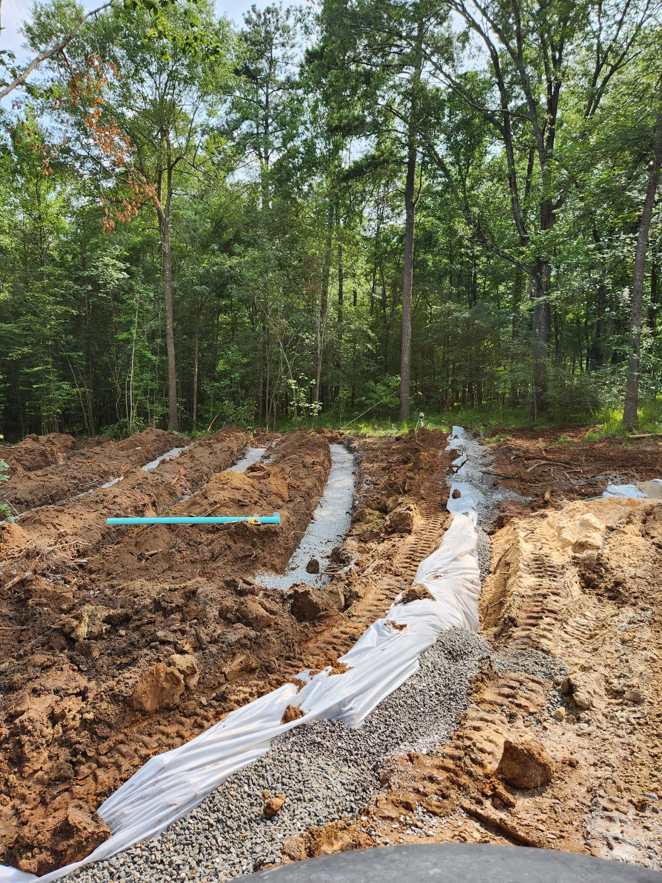 Construction site in a forested area with trenches filled with gravel and white geotextile fabric, and a blue pipe.