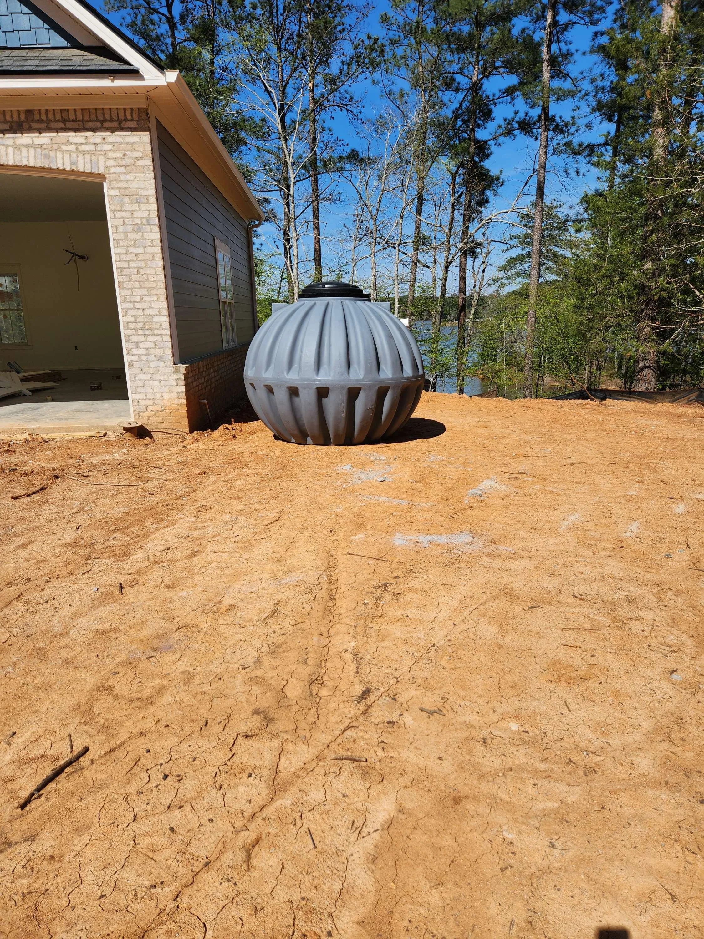 A large, gray, beside-ground rainwater collection tank or water storage tank outside a house surrounded by trees and dirt ground.
