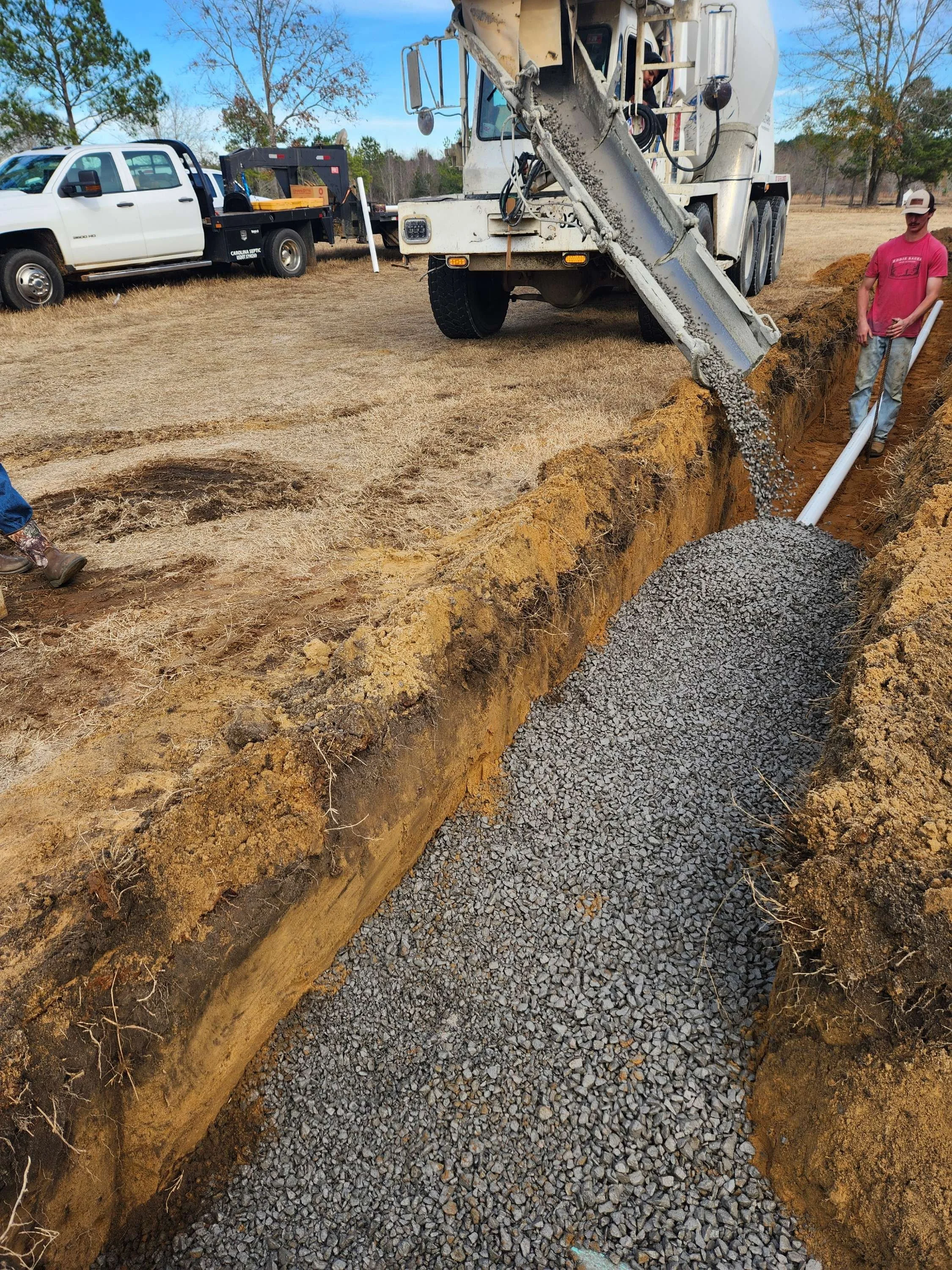 A construction site with a worker pouring gravel into a trench, with a cement mixer nearby and trucks in the background.