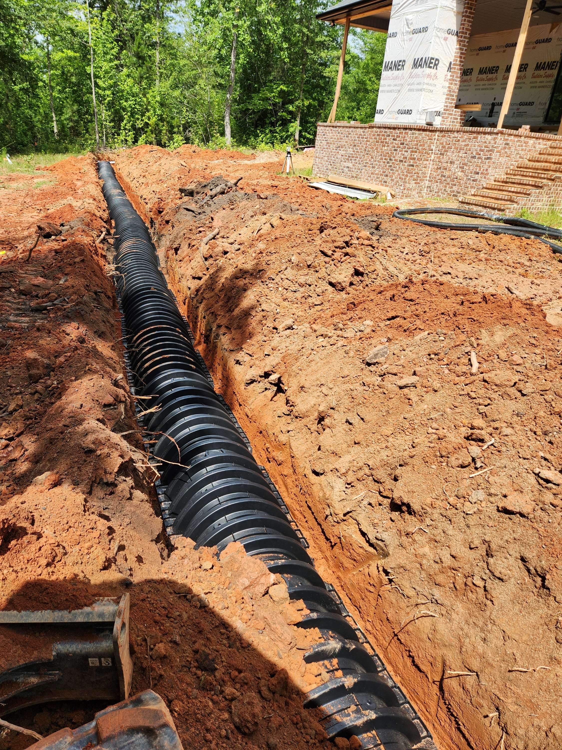Installing a black, corrugated drainage pipe in a deep trench near a house under construction, surrounded by reddish-brown soil and green trees.