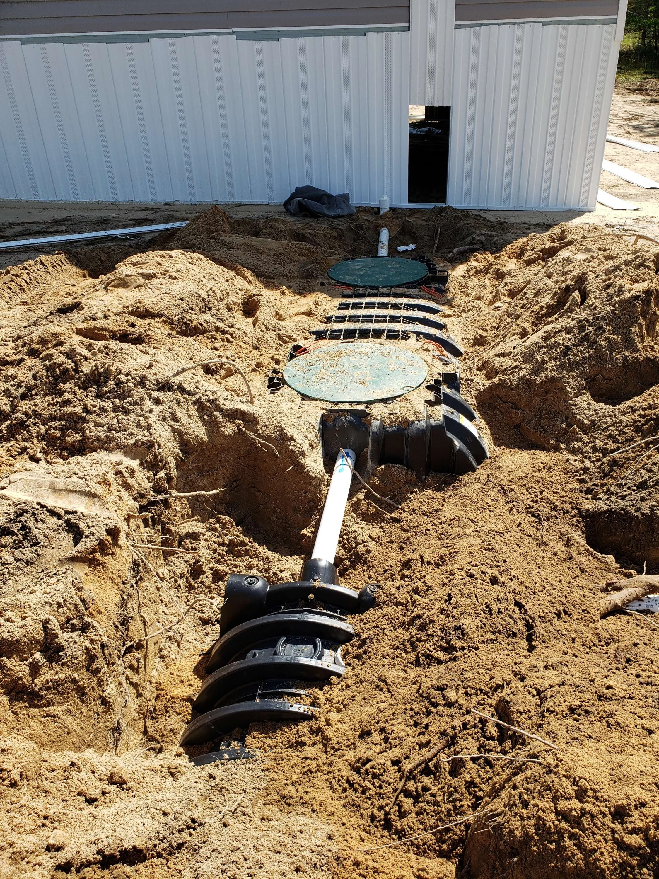 Underground utility access point with a pipe and black fitting, surrounded by dirt, in front of a metal building wall with a small opening.