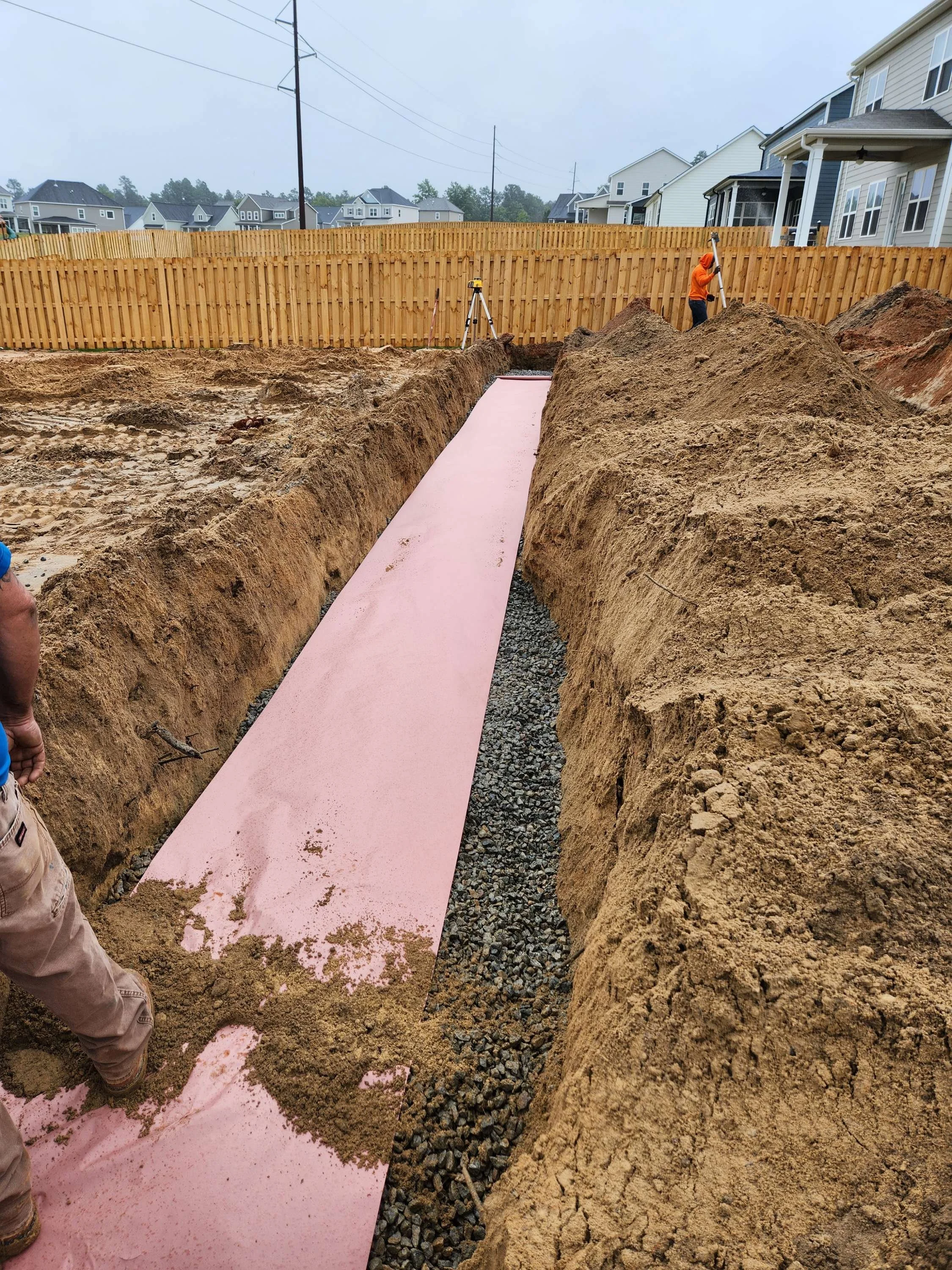 Construction workers installing a pink foam board insulation on a concrete slab foundation in a backyard, with a wooden fence and new houses in the background.