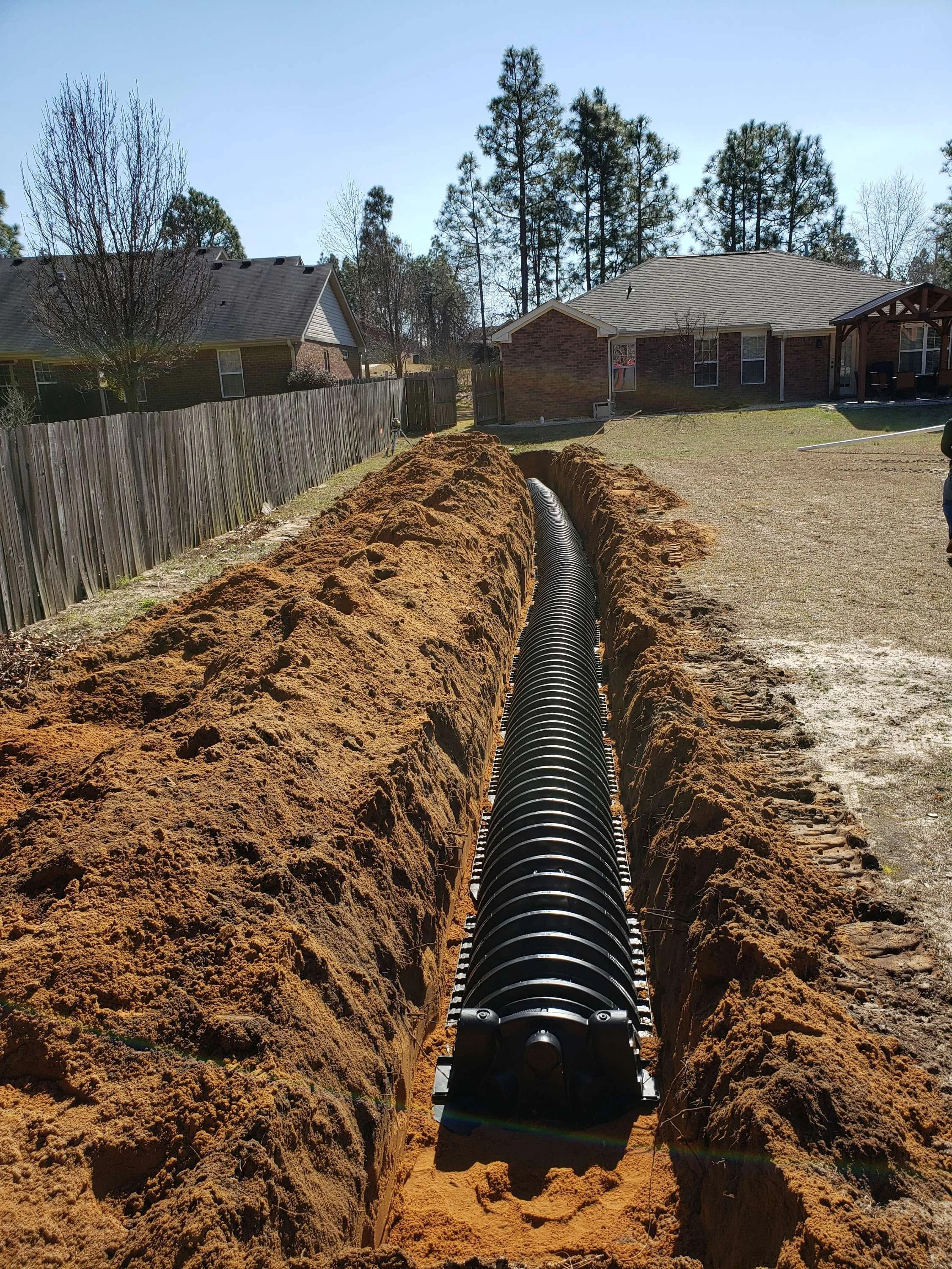 A long trench with black corrugated drainage pipe installed in the backyard of a suburban home during daylight.