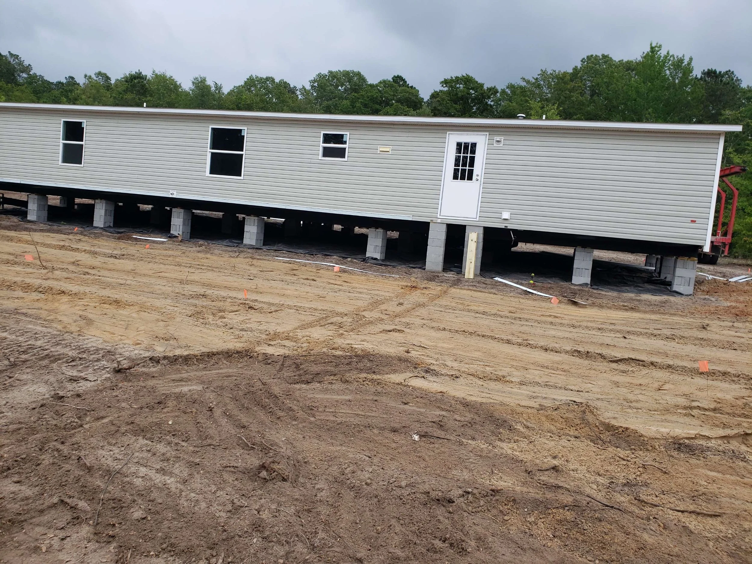 A long, light-colored mobile home on raised concrete blocks on a construction site with dirt ground and some scattered markers, surrounded by trees and overcast sky.