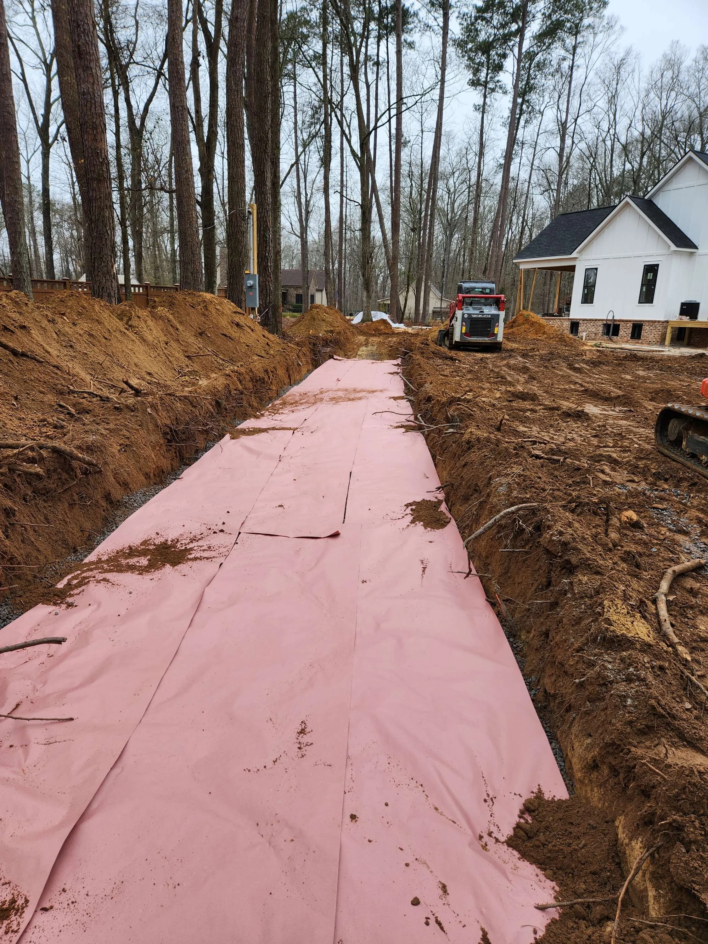 Construction site showing a trench with a pink vapor barrier laid down, surrounded by dirt mounds, with construction equipment and a partially built house in the background, in a wooded area.