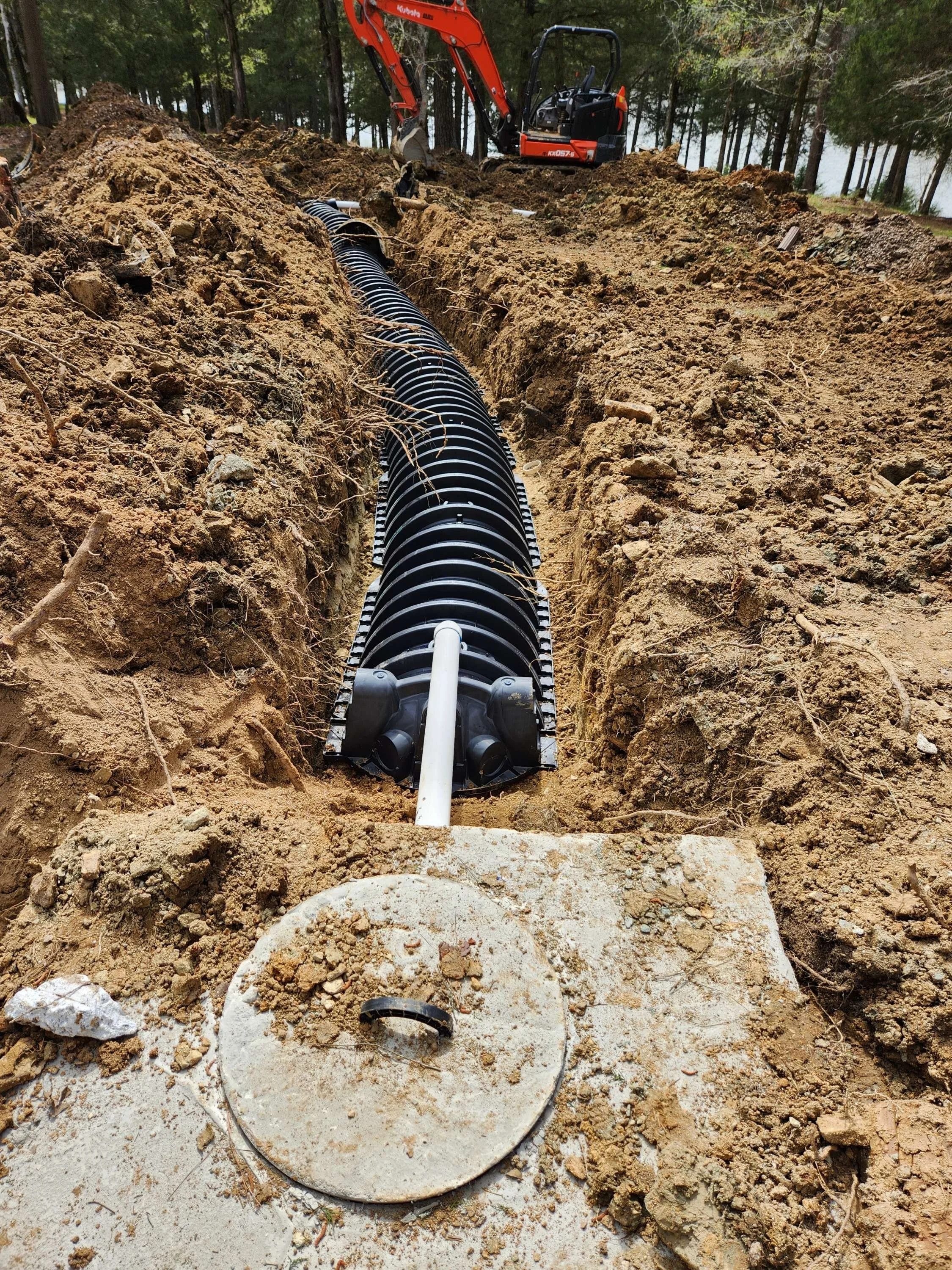 Installation of a large underground pipe in a trench dug in dirt, with construction equipment in the background.