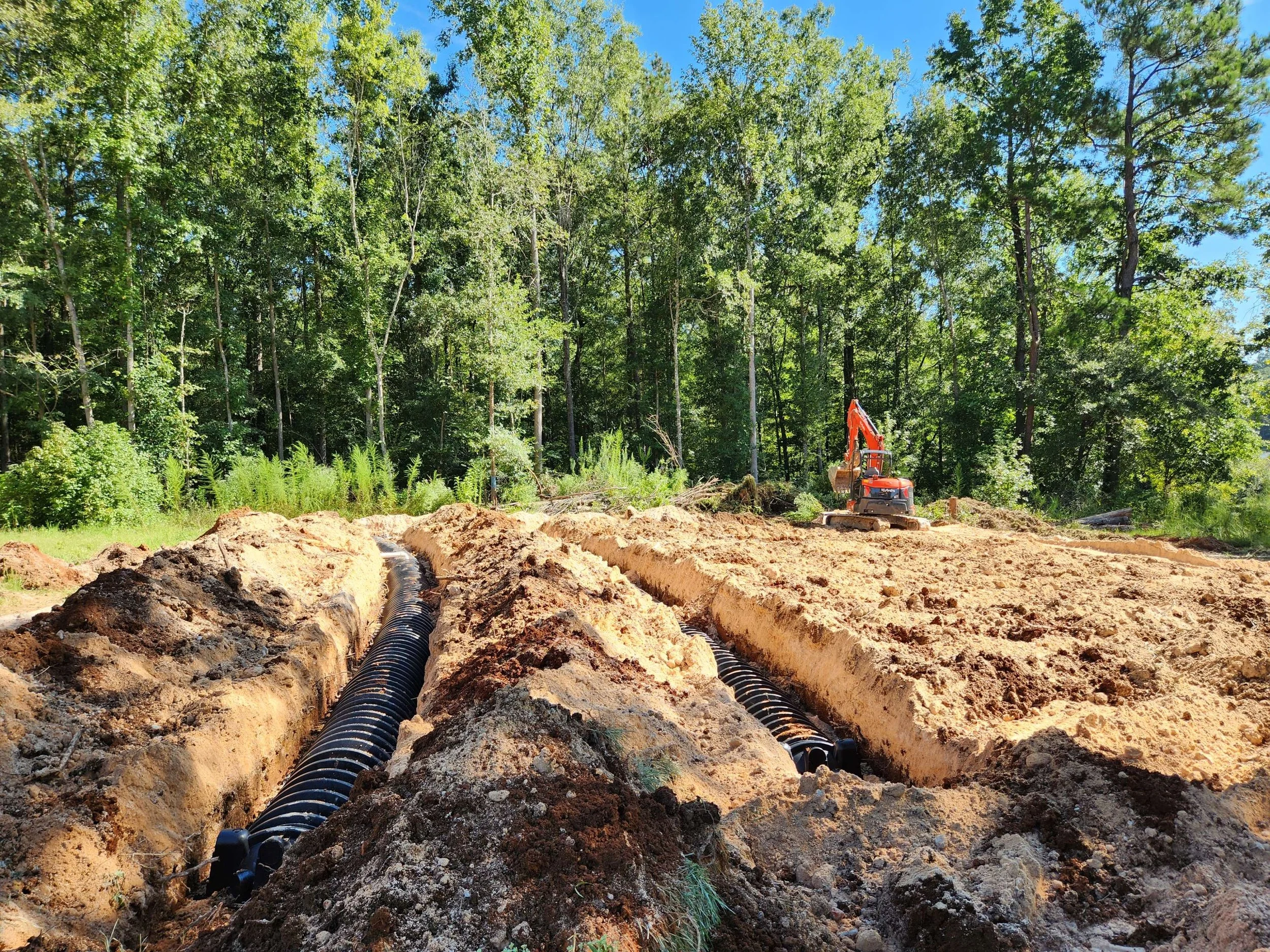 Construction site with trenches and black pipes being installed, surrounded by trees and a clear blue sky, with an orange excavator in the background.