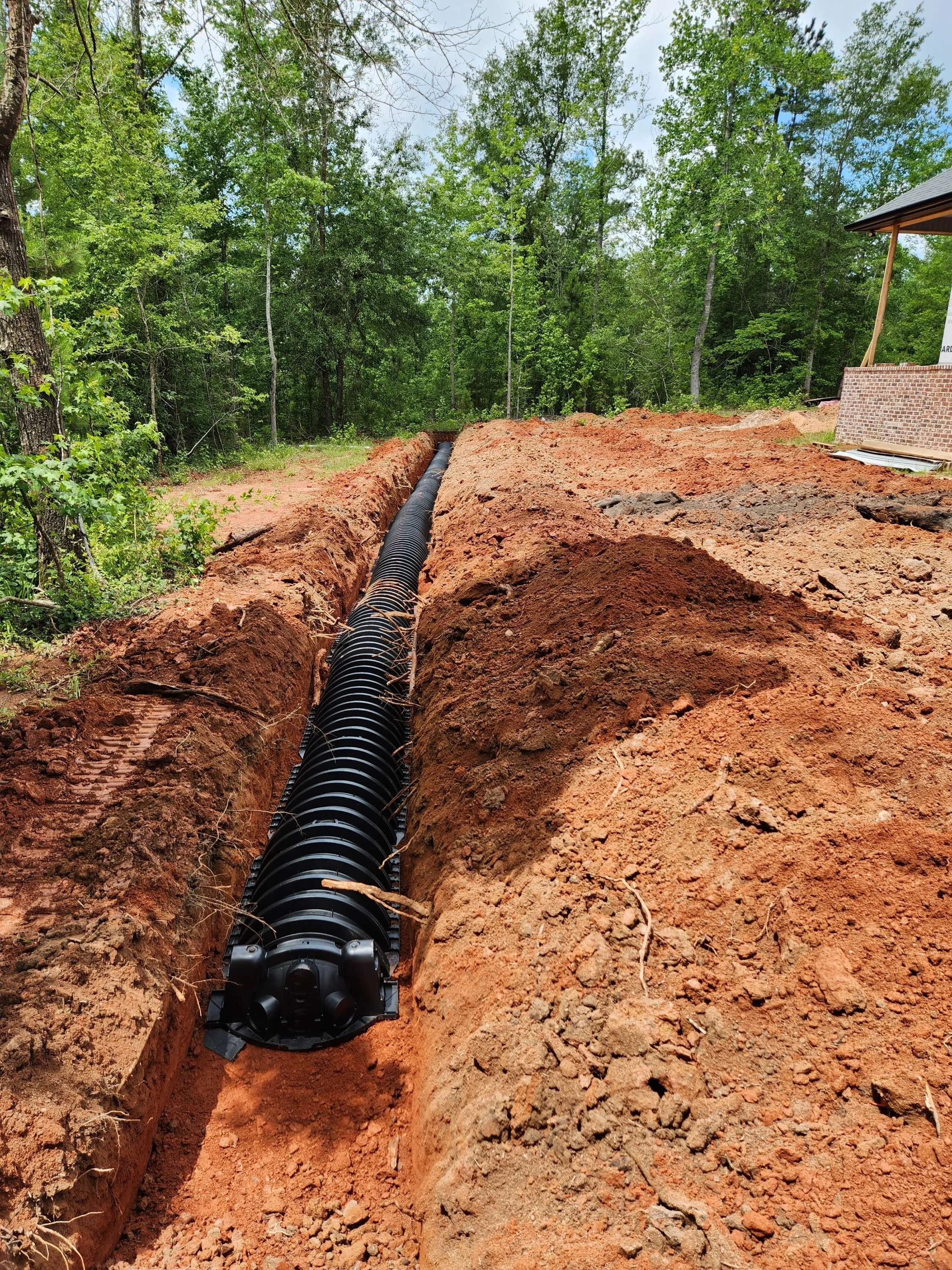 A black corrugated pipe is being installed in a trench in a wooded area.