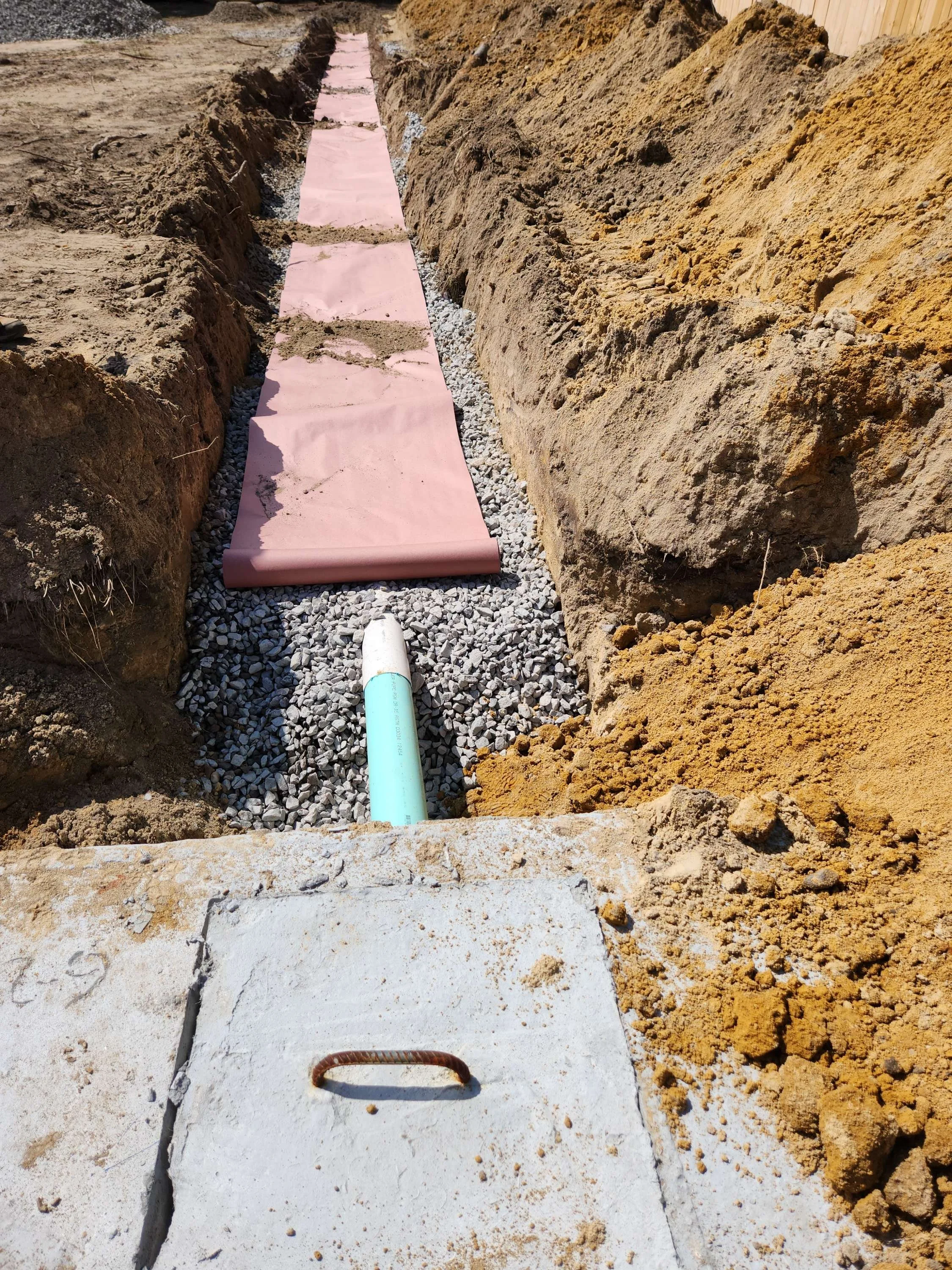 Construction site with a trench, pink insulation foam boards, gravel bed, PVC pipe, concrete curb, and dirt walls.