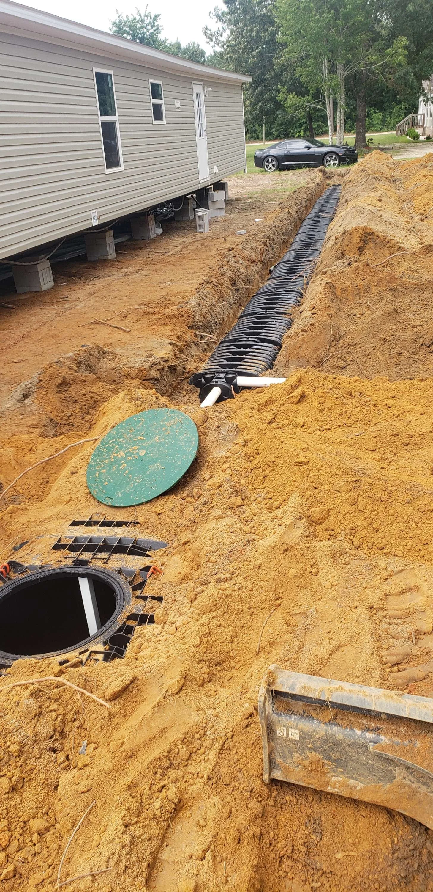 Underground utility installation with black pipe or conduit running through a dug trench beside a beige mobile home with a parked black car nearby.