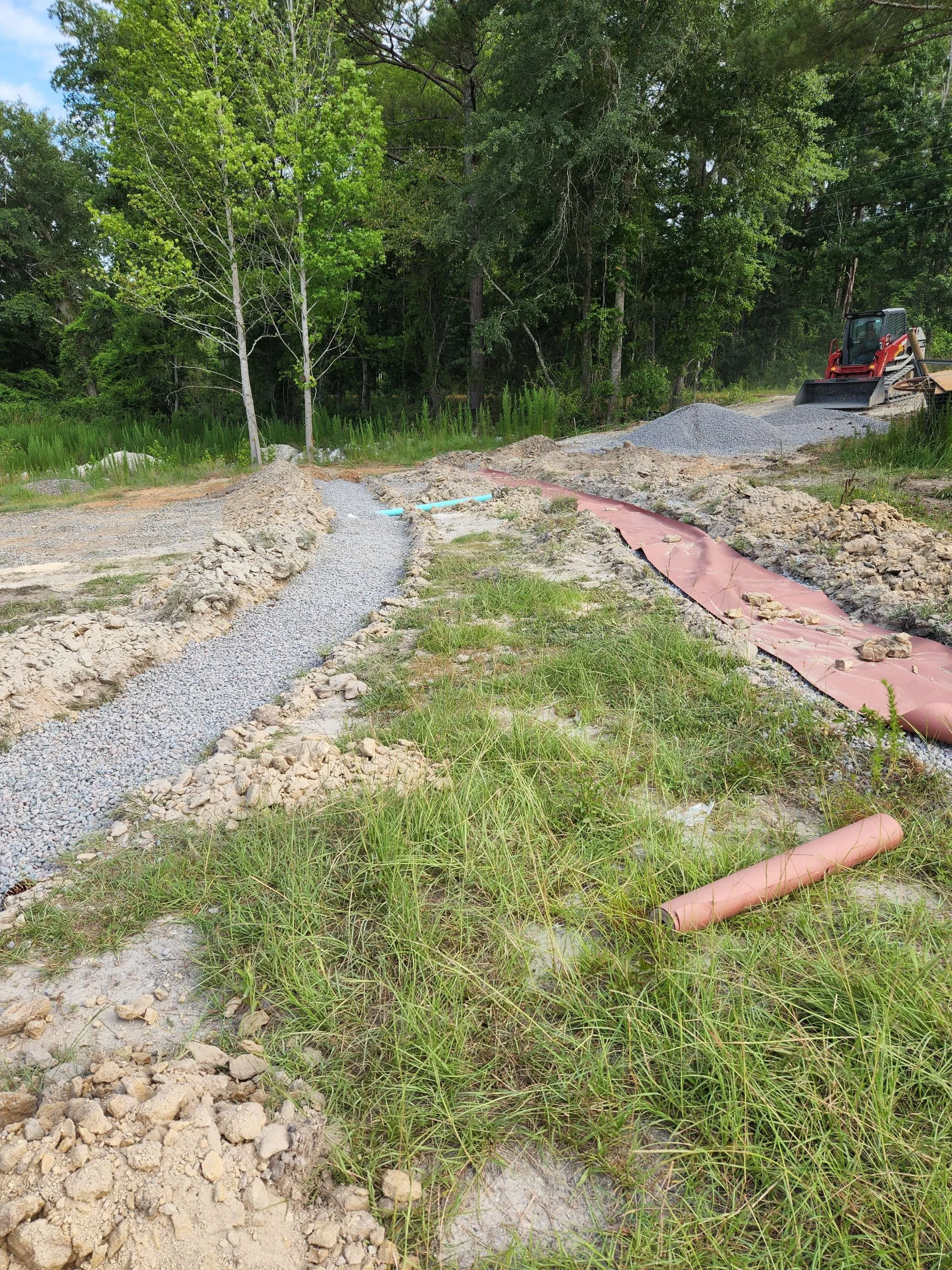Construction site with narrow gravel pathway being laid in a natural area with trees in the background, a red construction vehicle, and pink drainage pipes.