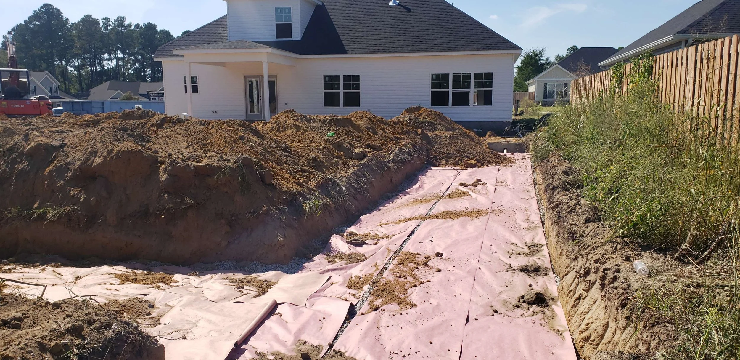 Construction site with dug trench and pink fabric ground covering in a residential backyard, with a white house and neighboring houses in the background.