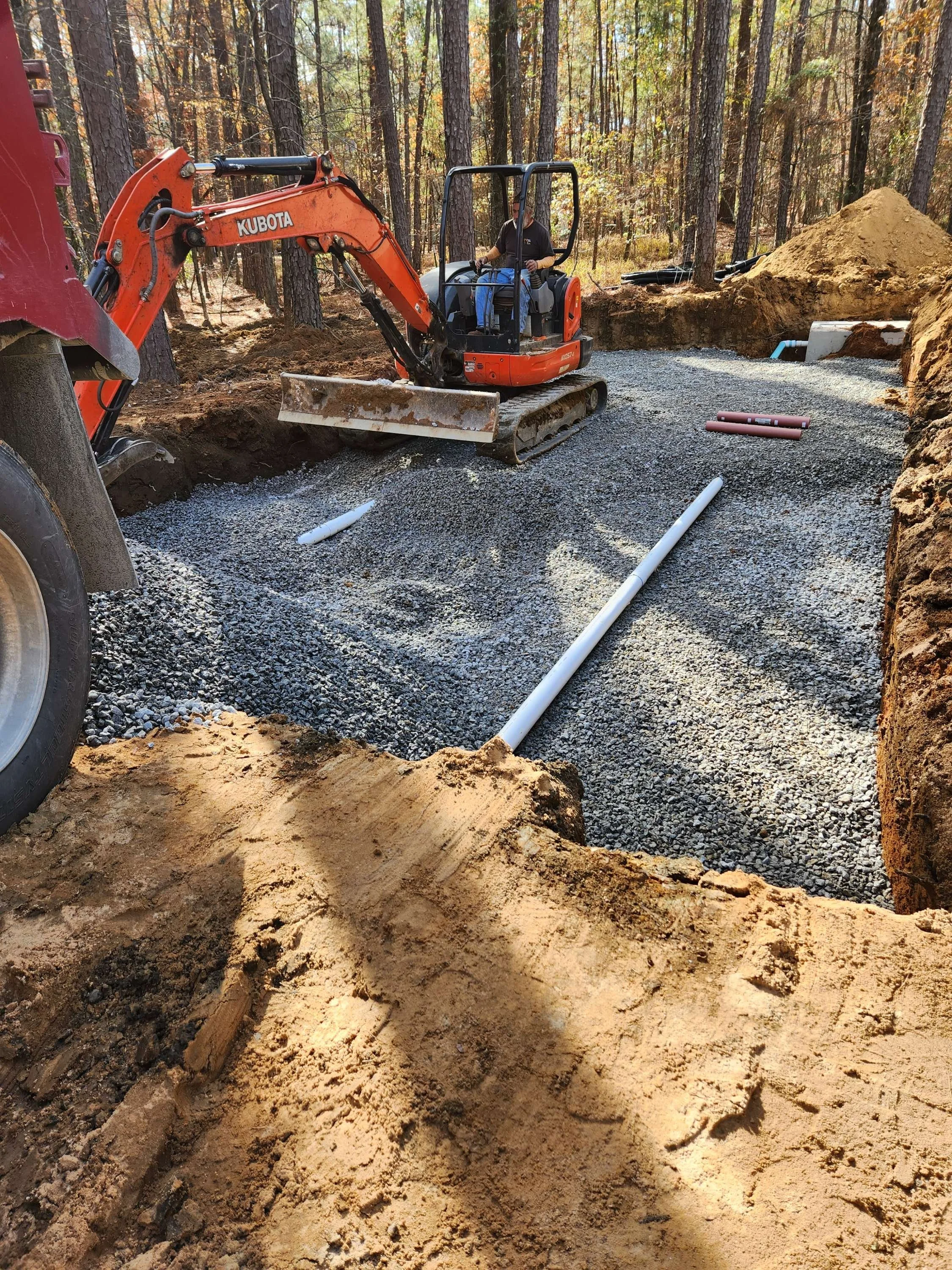 A construction site in a wooded area where a man is operating an orange Kubota mini excavator, surrounded by dirt and gravel pipes being laid for future construction.