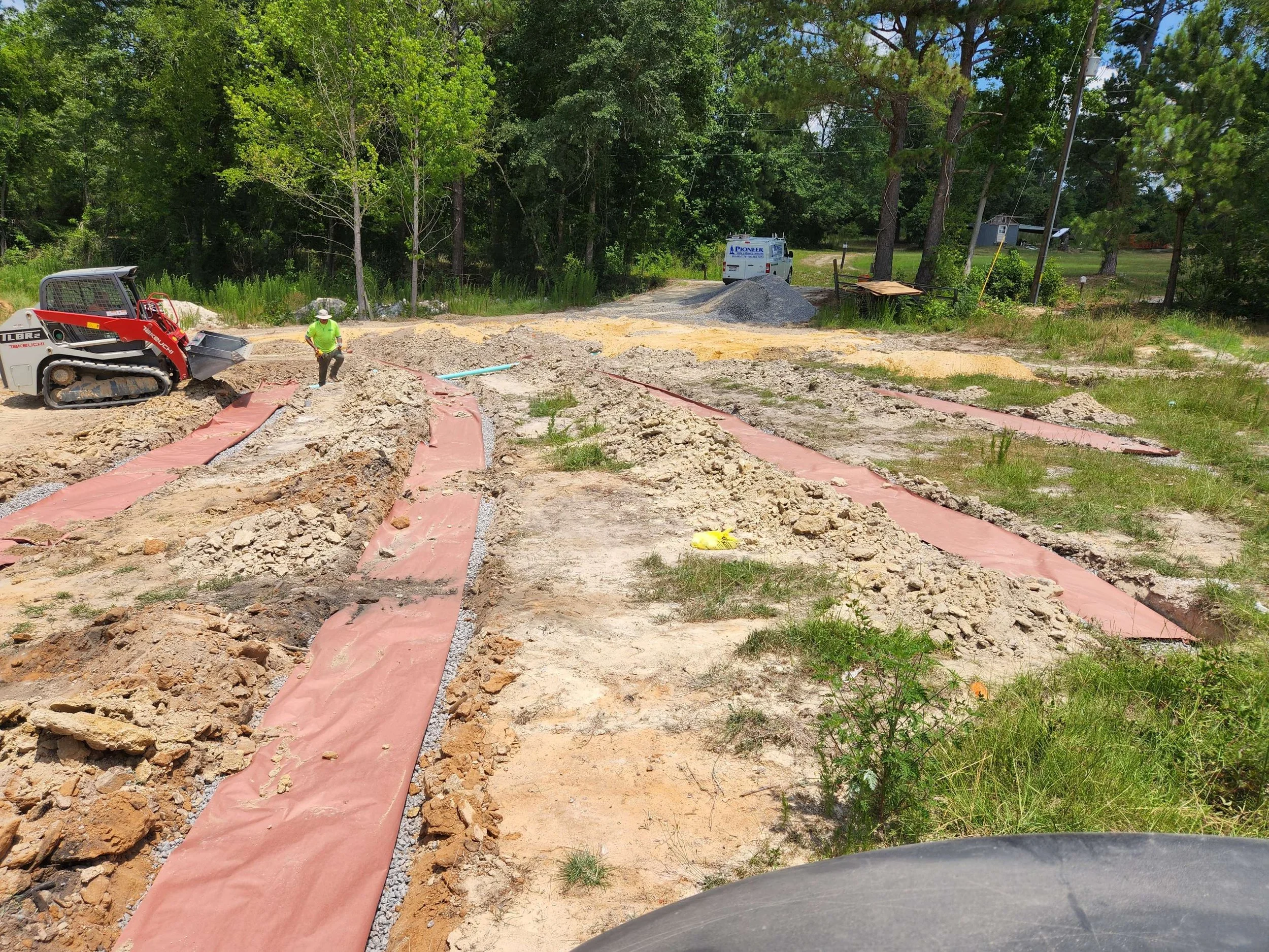 Construction site with dirt, pink sheathing, and gravel, a worker in neon green safety vest and white helmet, a small tracked excavator, and vehicles in the background surrounded by trees.