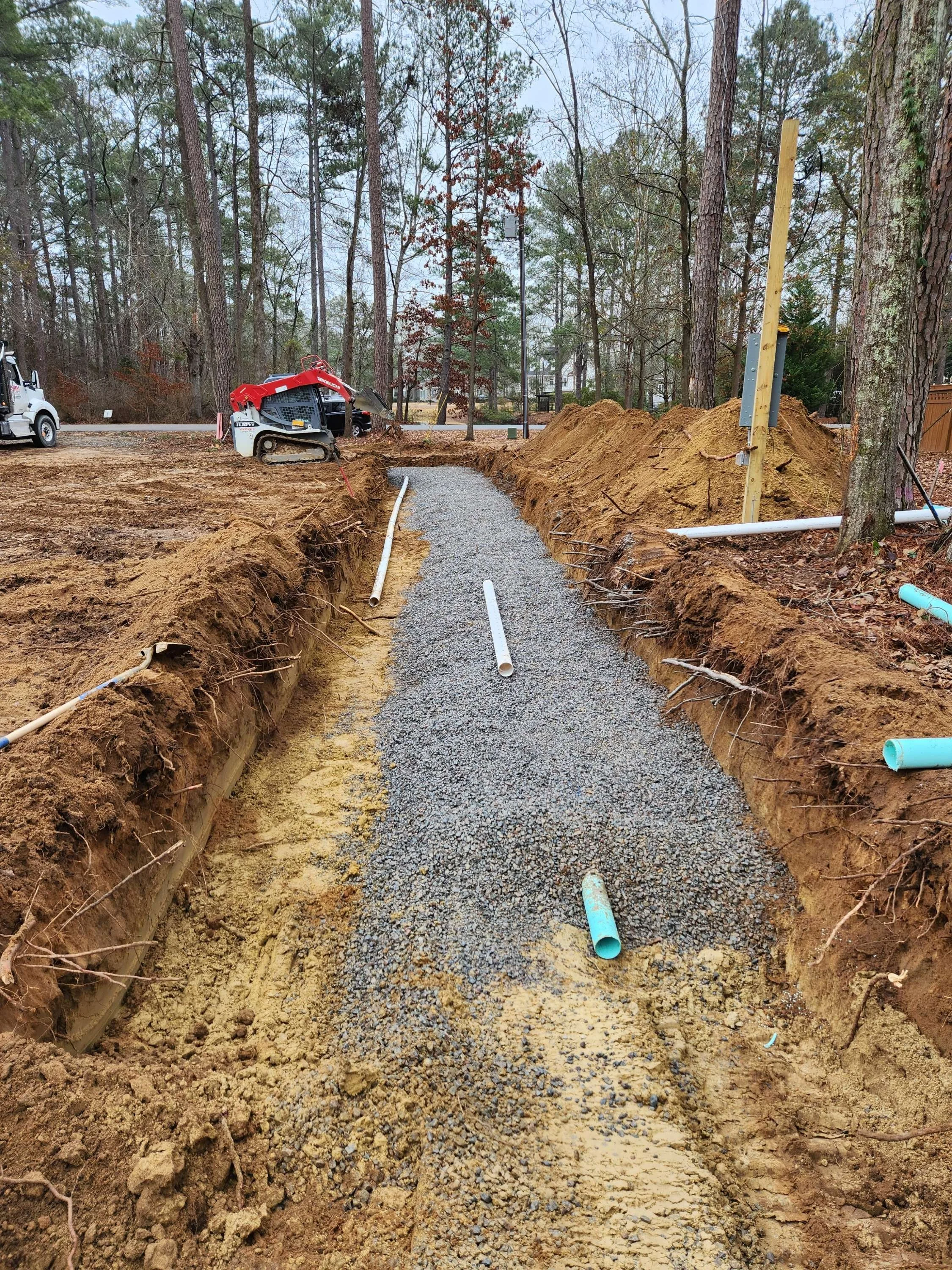 Construction site with a gravel pathway, PVC pipes, and a small tracked excavator among trees, with dirt piles on either side.