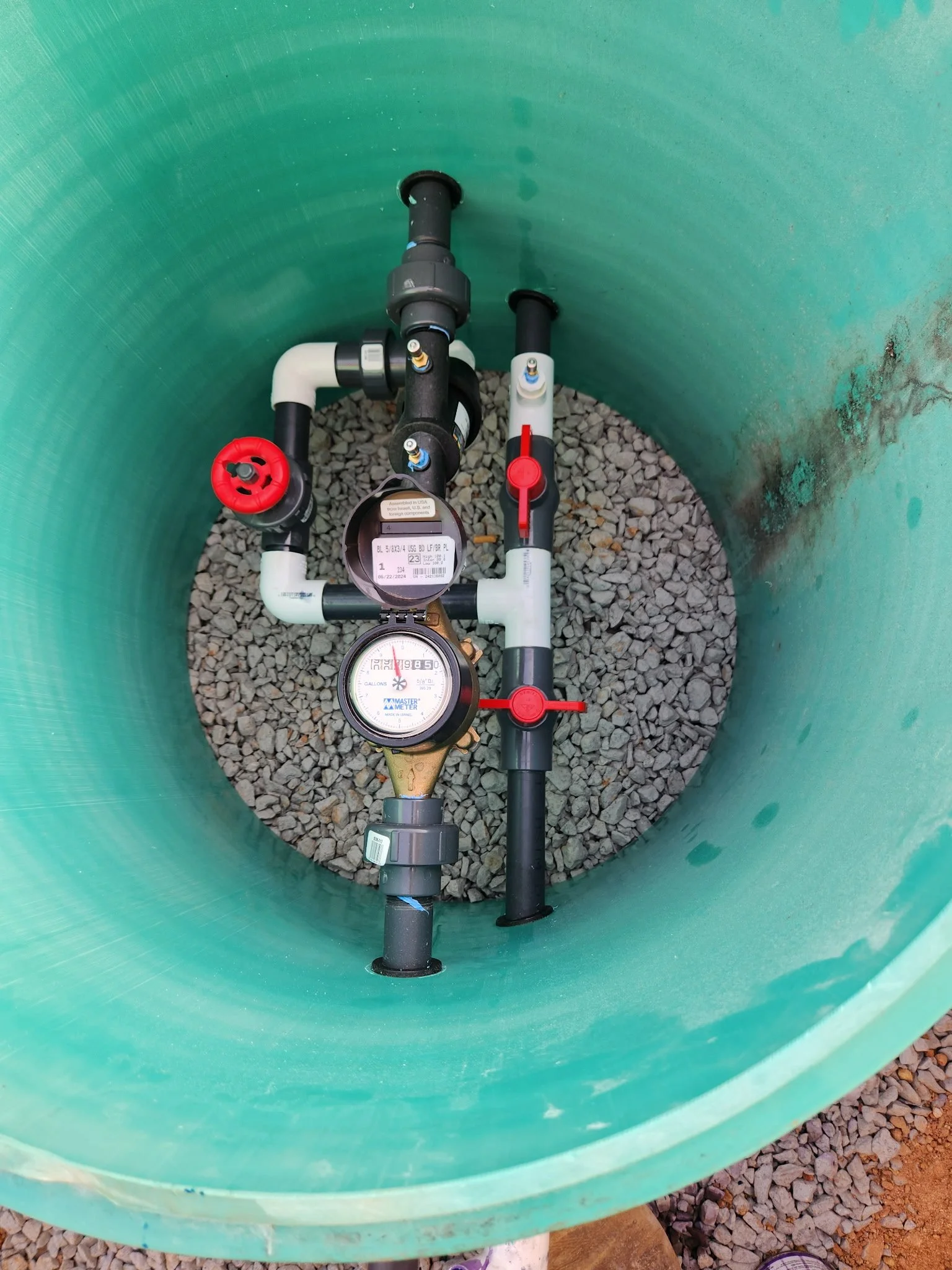 Inside a green cylindrical water tank, there are black and white plastic pipes, a water meter, and red valves, with gravel at the bottom.