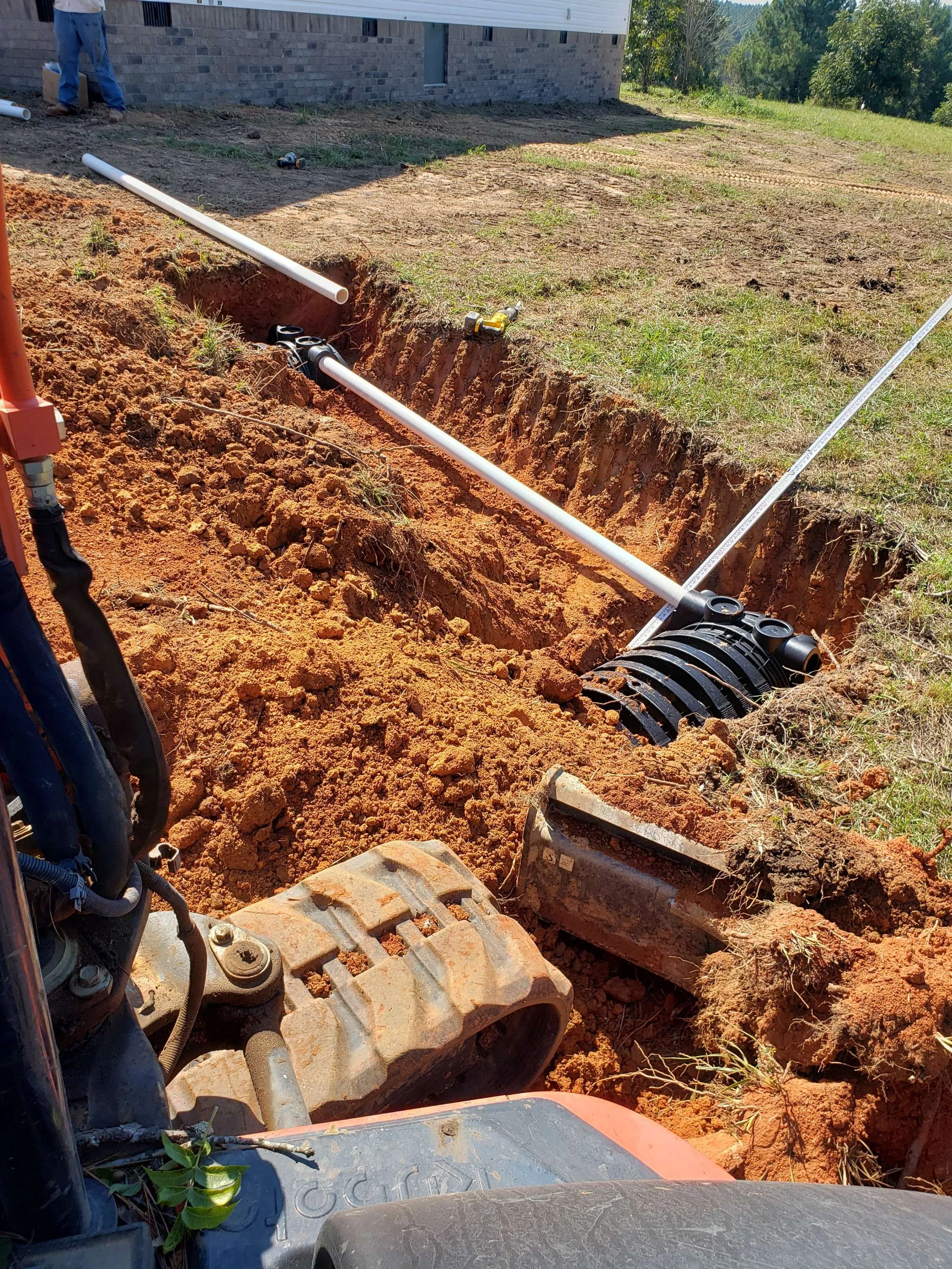 Excavation site with a trench for underground piping, showing a bulldozer or excavator with pipes and fittings.