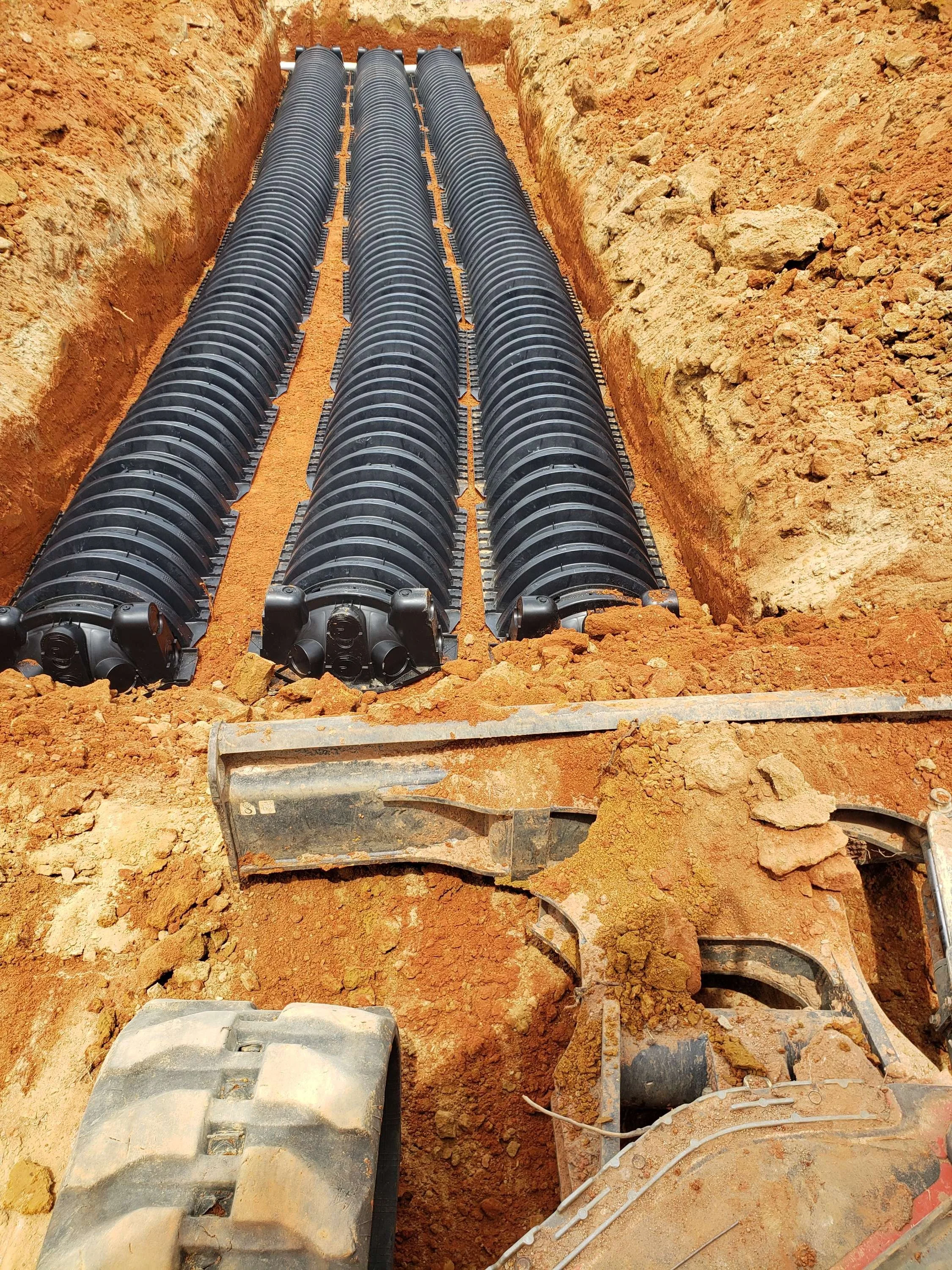 Underground pipe installation with three black corrugated pipes in a trench, with construction machinery parts in the foreground.