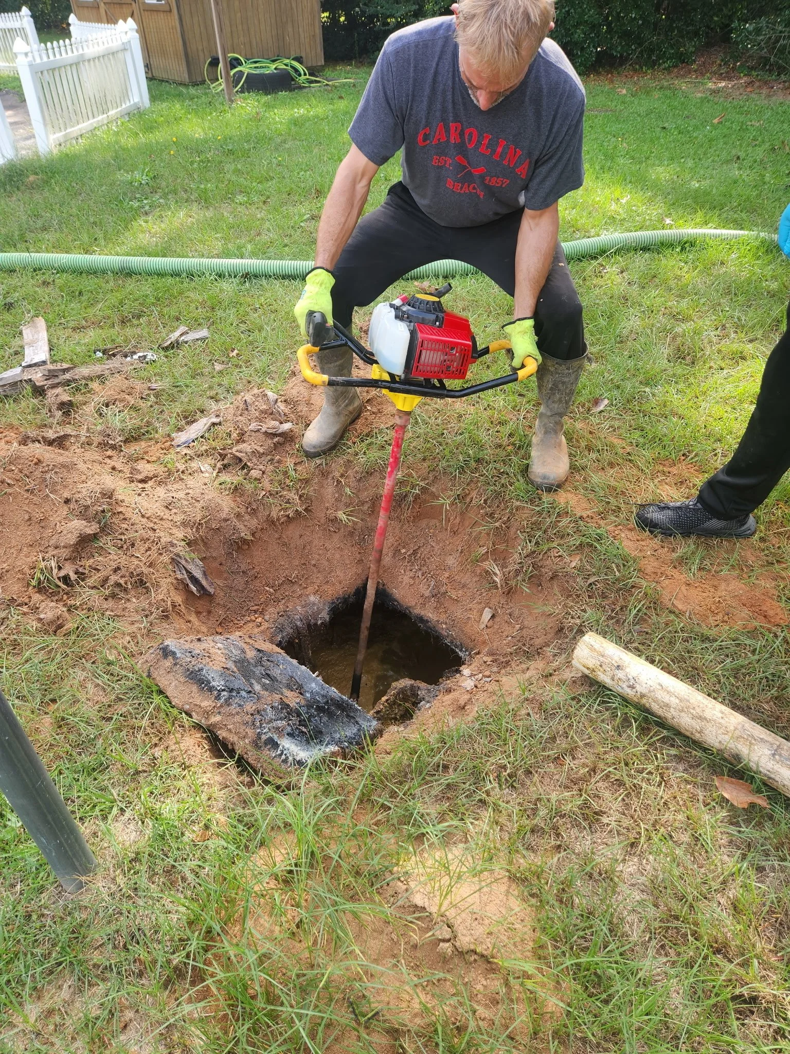 Person using a power auger to drill into the ground in a grassy yard, wearing a gray Carolina Beach T-shirt, gloves, and boots.